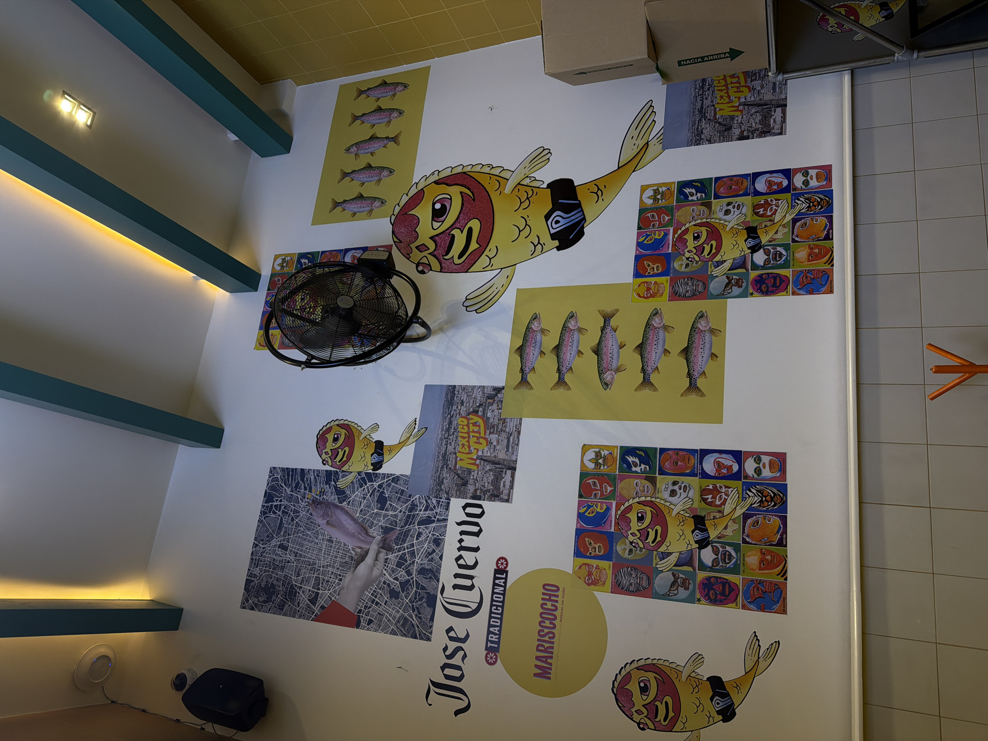 The image shows a wall in a restaurant or business in Mexico City. The wall is white with various decals and posters. Some of the notable decorations include:
* Decals of fish with Lucha Libre masks.
* Posters of fish in rows.
* A poster of Mexico City.
* A poster with the words "Jose Cuervo Tradicional Marisococho" in large print.
* A poster featuring a hand holding a fish superimposed over a city map.
A black stand-up fan is affixed to the wall. The location appears to be indoors, and possibly a casual dining establishment.