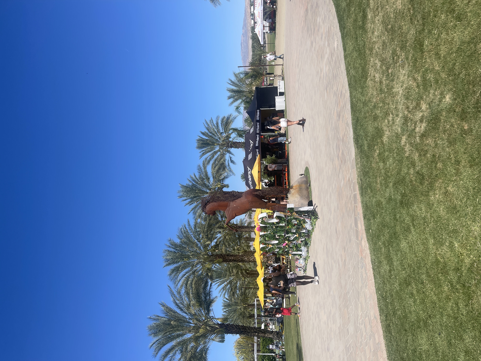 A partly sunny outdoor scene in Thermal, United States, with a clear blue sky and numerous tall palm trees is depicted. The scene includes a bronze-colored horse sculpture, vendor stands, and various decorative elements, including yellow umbrellas and floral arrangements. People are present, either sitting or standing near the vendor stands. The ground comprises a brick pathway and a green grassy area. In the background, a mountain range is visible, alongside some buildings.