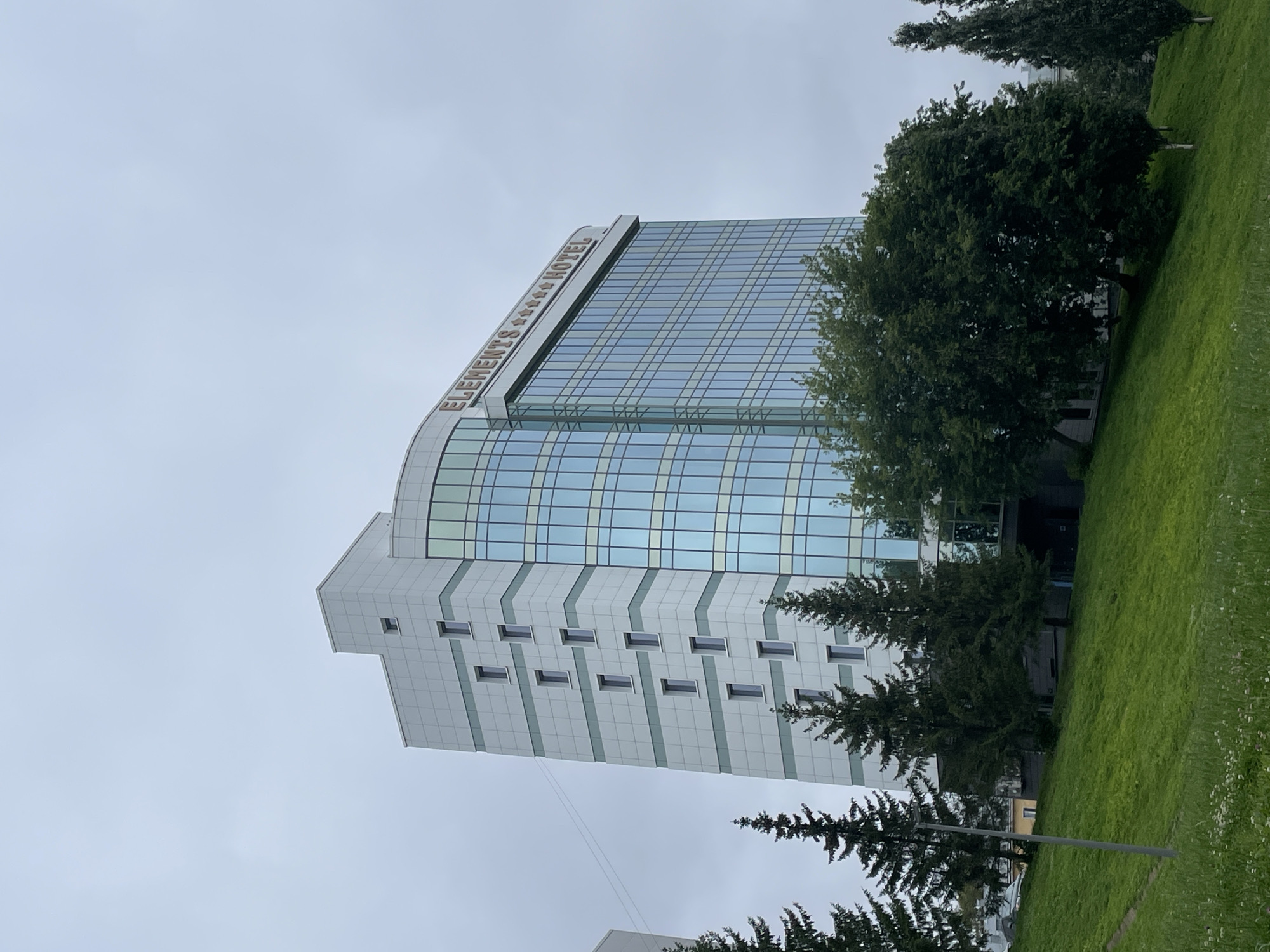 The image shows a tall, modern hotel building with a glass facade, identified by signage as "ELEMENTS **** HOTEL." The building is located in the city of Kirov, Russia. In front of the hotel is a grassy slope, dotted with a few wildflowers. Several evergreen trees, possibly fir or spruce, are strategically placed along the slope and in front of the hotel's lower levels. The sky is overcast, indicating a cloudy day. There are no people visible in the image. The overall scene depicts an urban landscape with a focus on the hotel and its surrounding greenery.