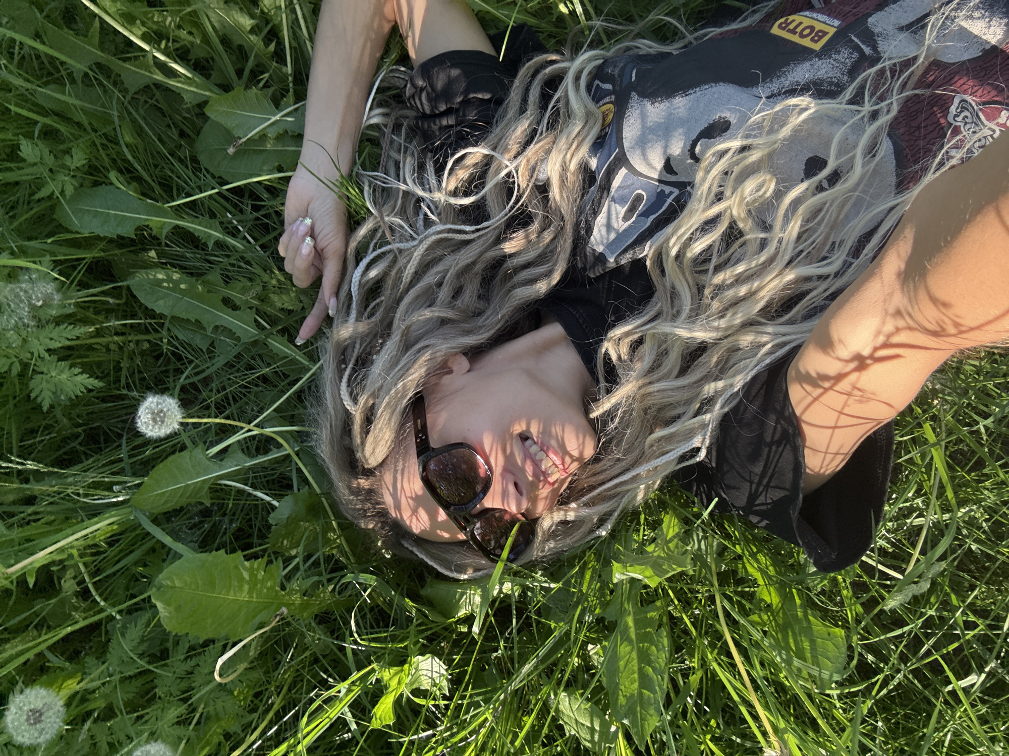 The image depicts an adult individual, appearing female, lying supine in a dense patch of sunlit green grass located in Rusanovo, Russia. The subject's head is tilted upwards, looking towards the sky. She wears dark sunglasses and has long, wavy blonde or light brown hair interwoven with lighter, possibly braided, strands, spread out on the grass. Her mouth is slightly open, revealing teeth. She is dressed in a black short-sleeved t-shirt featuring a prominent graphic design on the front, with a yellow rectangular label reading "BOTR" in black text visible on the lower left side of the shirt. Her left arm (viewer's perspective) is bent with the hand resting near her head, while her right arm (viewer's perspective) extends slightly outwards, fingers lightly curled. Her fingernails are manicured with a light, possibly glittery, polish. Distinct shadows from overhead foliage are cast across her skin and clothing. The grassy environment includes various broad-leafed plants and several white, spherical dandelion seed heads.