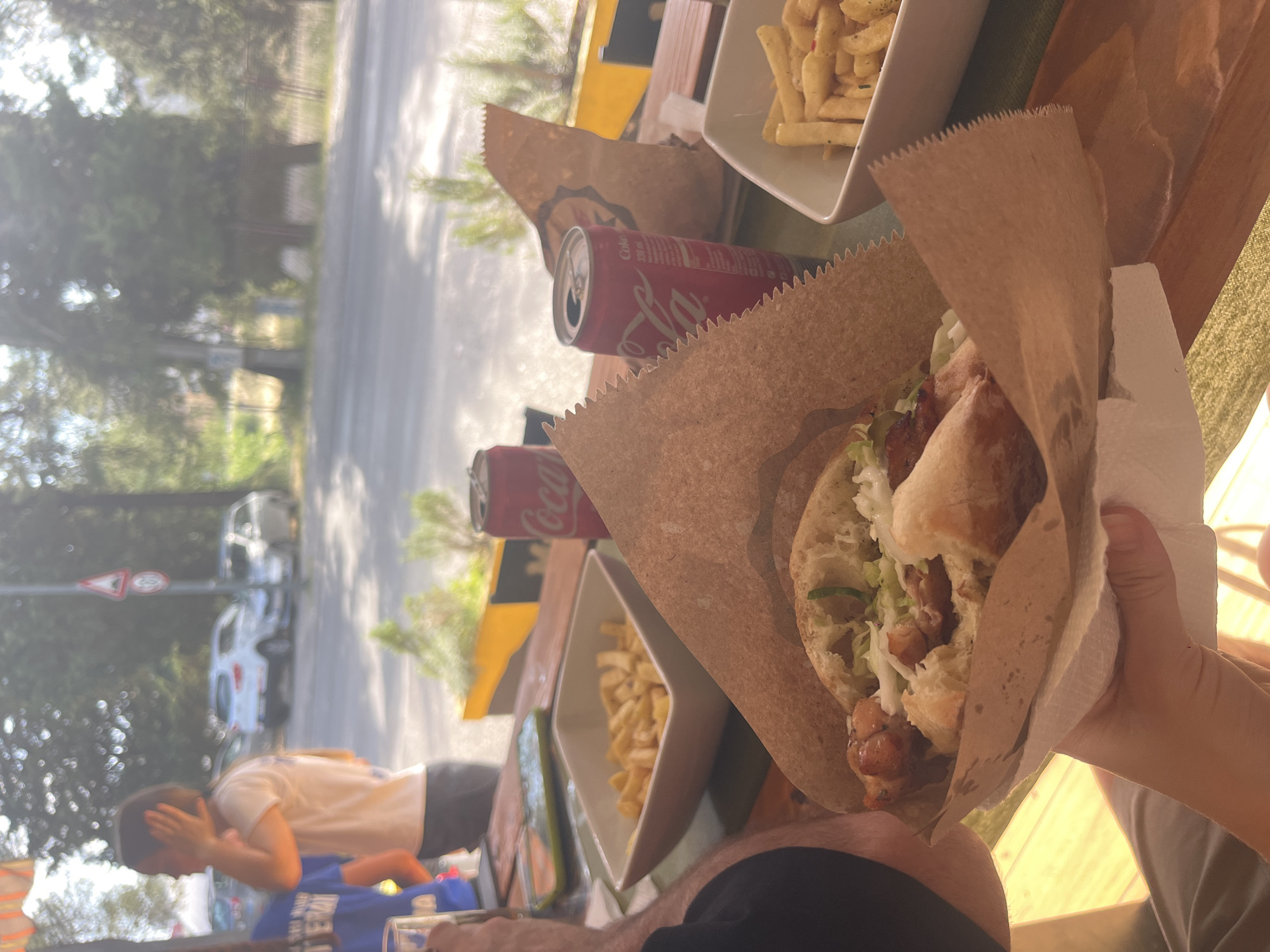 The image shows a partially eaten kebab wrapped in brown paper held in a hand. Next to the kebab are a can of Coca-Cola, a dish of fries, and another kebab wrapped in paper. In the background, there is a street scene with trees, cars, and a pedestrian crossing sign. A person is standing by the street with their head in their hand. The location is likely in Podgorica, Montenegro.