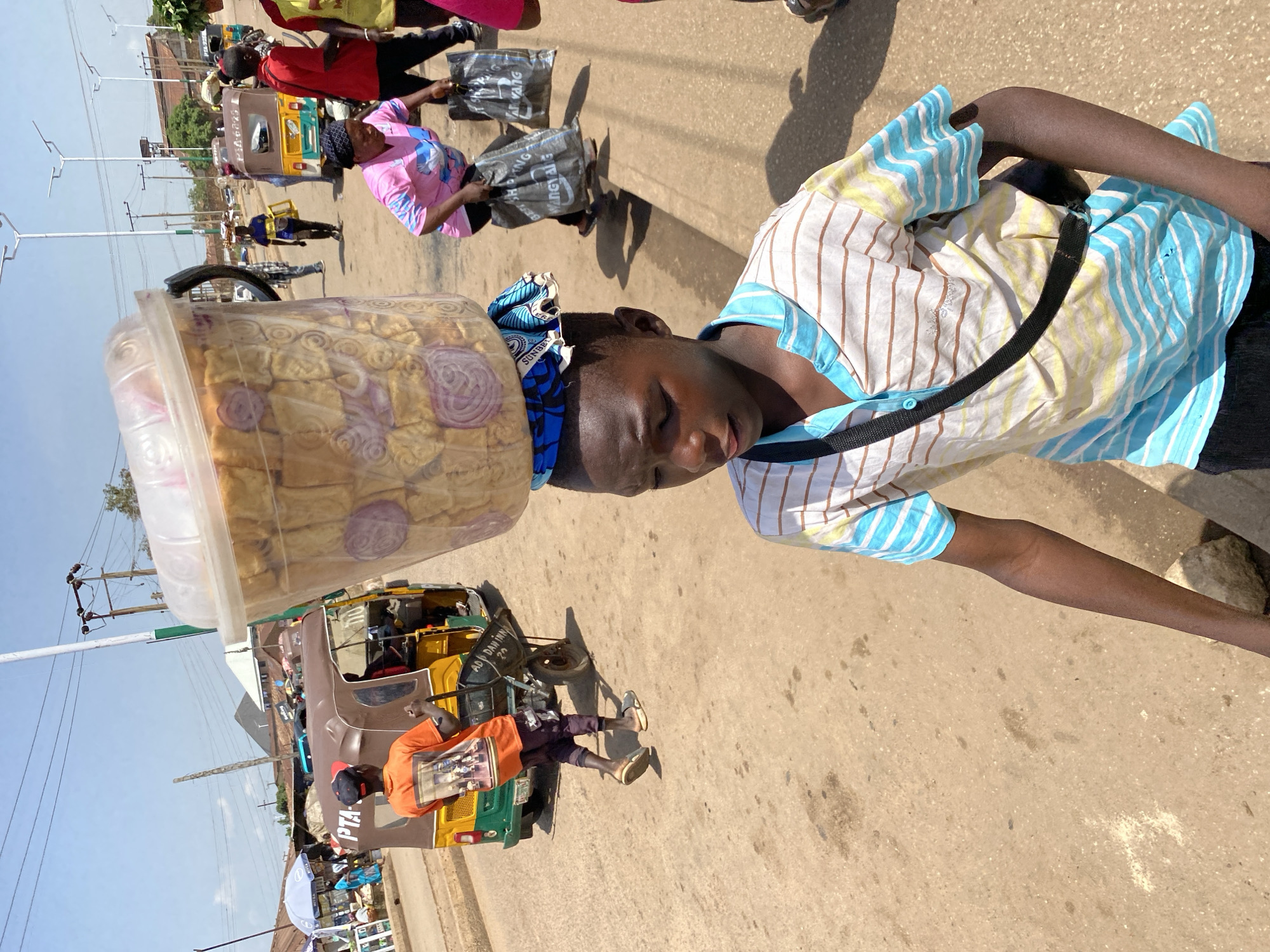 In Bukuru, Nigeria, a young Black boy walks forward, carrying a large, clear plastic container filled with food items, balanced on his head. The container is covered with a blue and white patterned cloth. He wears a striped short-sleeved shirt and dark pants, with a black strap across his chest.
The environment is a sun-drenched, dusty urban street. In the background, a colorful auto-rickshaw, a common mode of transport, is visible. Other people are present, including a woman in a pink shirt carrying shopping bags and a person in an orange shirt pushing a cart. The scene depicts a bustling market or street scene.