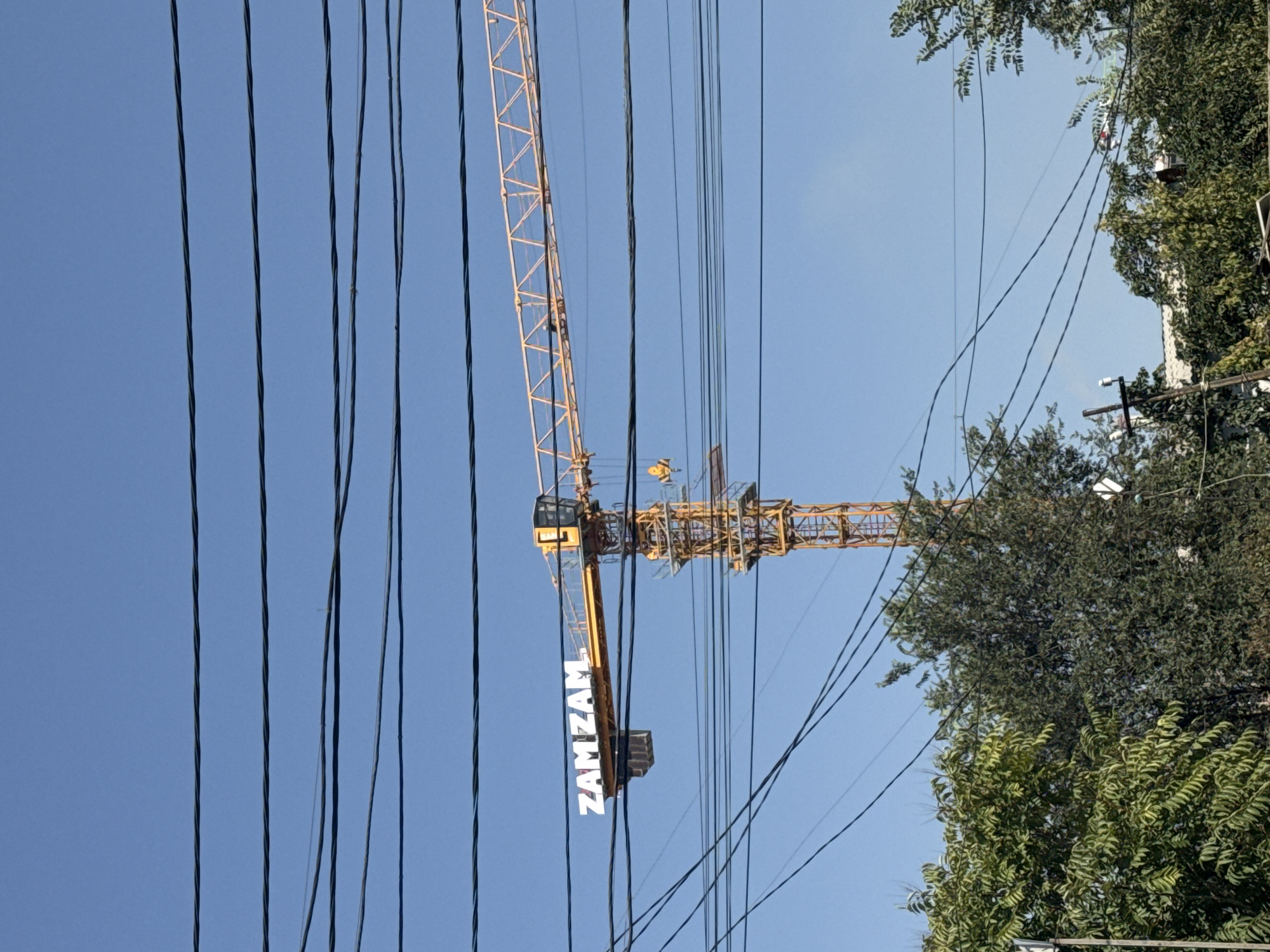 The image captures a bright, sunny day with a clear blue sky, partially obscured by several thick black electrical wires stretched across the frame. Dominating the mid-ground is a tall, yellow construction crane. The crane has a prominent horizontal boom, on which the white, capitalized letters spelling "ZAMZAM" are clearly visible. Below the boom, the crane's central tower structure is intricate and made of metal supports, with a small cabin at the top of the tower. A yellow hook and other equipment are attached to the crane's boom and tower. In the foreground, the tops of lush green trees are visible, suggesting an urban environment, possibly in Bishkek City, Kyrgyzstan, with some smaller trees and structures interspersed amongst them. The overall scene suggests an active construction site within a developed area.