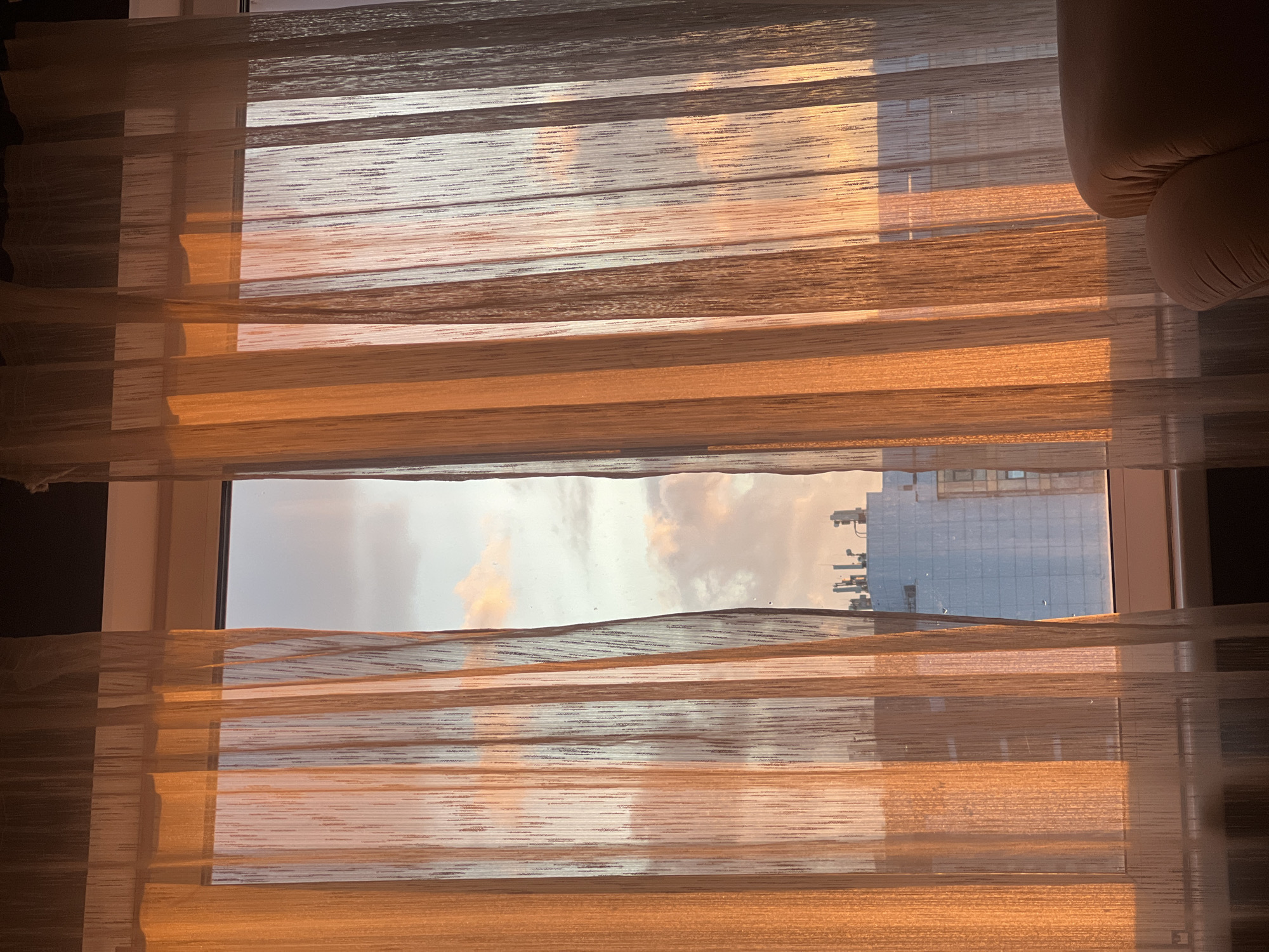The image shows a window with semi-transparent blinds, partially obscuring the view outside. Through the blinds, one can see a cloudy sky with a hint of sunset colors and a building in the distance. The building appears to have a grid-like pattern of windows. The blinds are horizontally striped in shades of tan and white. The blinds are slightly open, allowing some light and the view to be visible. Part of a couch or chair is visible on the right side of the image. The setting is a room in a building, and the view suggests an urban environment. There is no explicit confirmation this is Krasnoyarsk, Russia.