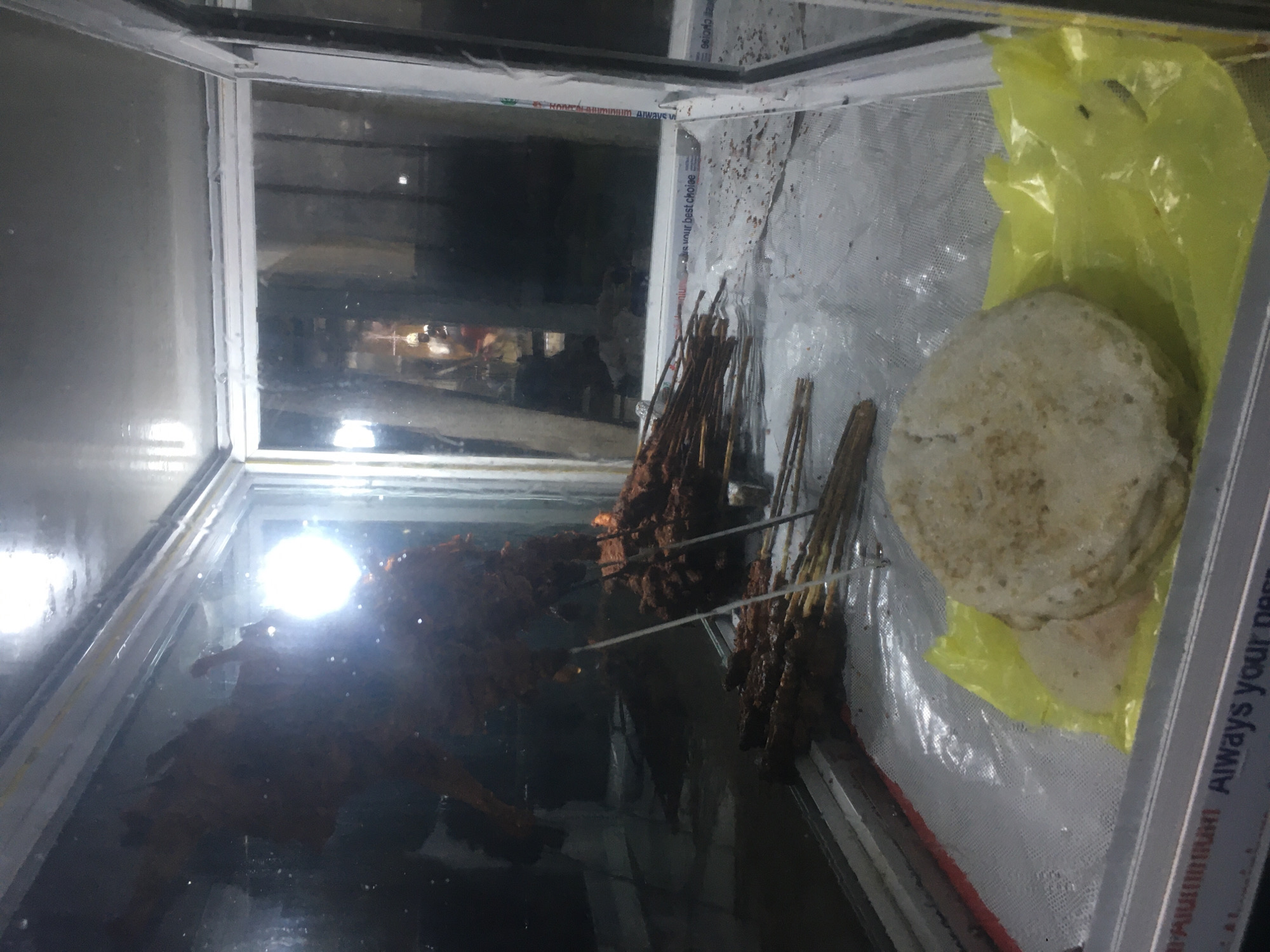 In a display case, likely a food warmer, possibly located in Gwarinpa, Nigeria, are several skewers of cooked meat, possibly suya, stacked on the left and in the middle. To the right, a stack of flatbreads (possibly roti or chapati) are partially wrapped in a yellow plastic bag. The display case appears to be in a public area, as indicated by blurred figures and a lighted interior reflecting off the glass. The case is made of metal and glass with condensation on the glass panes.