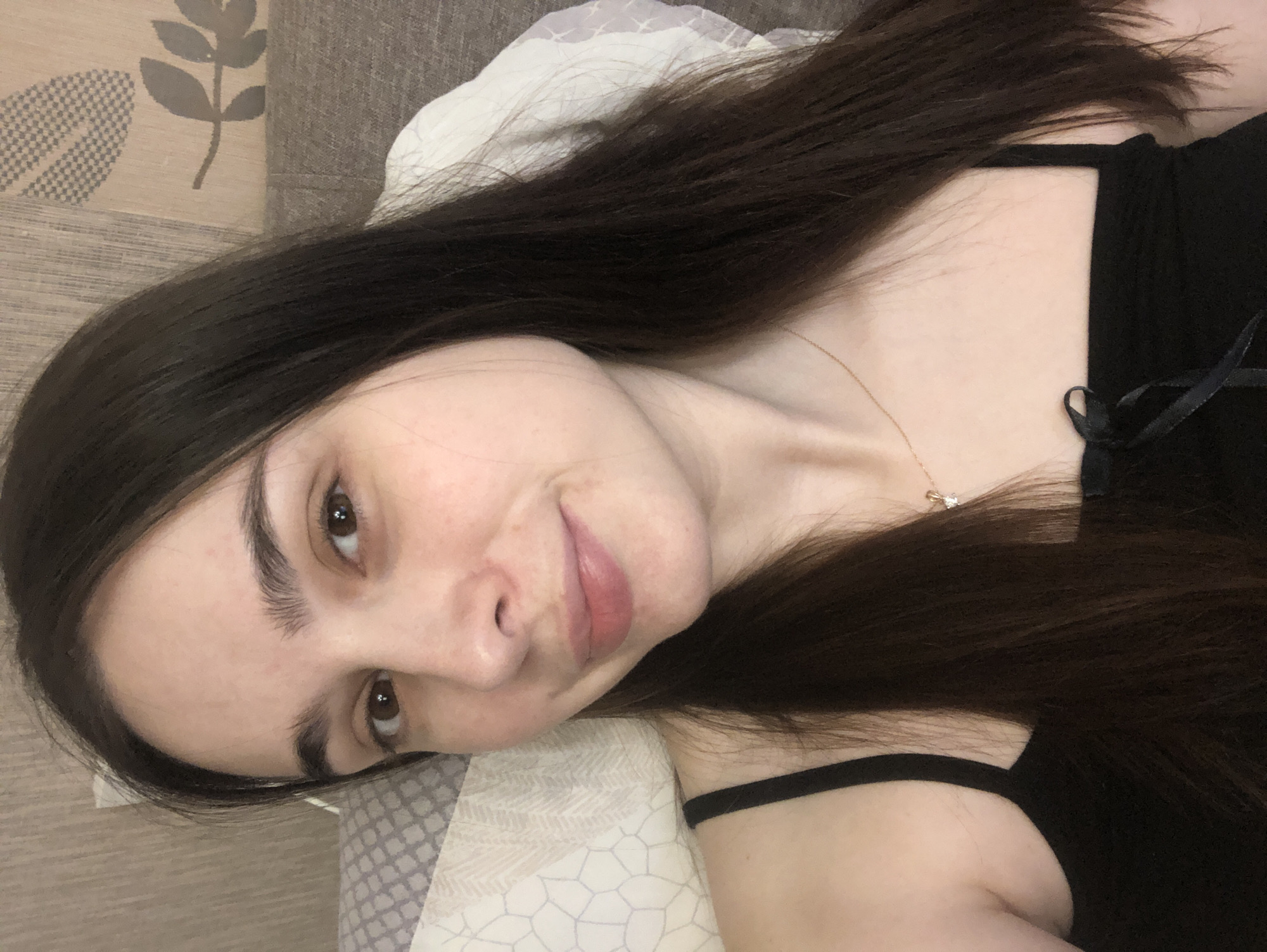 A single female subject is depicted in a close-up, chest-up shot, ostensibly within an indoor setting in Chaikovsky, Russia. The subject has fair skin, long, straight, dark brown hair that extends over her shoulders, and brown eyes. Her face is directed towards the camera with a slight, closed-mouth smile. She is wearing a black spaghetti-strap top, which features a small black bow tied at the center of the neckline. A delicate gold-colored chain necklace with a small, clear pendant is visible on her neck and upper chest. The immediate background to the subject's left includes a light-colored pillow with a geometric pattern. To her right and behind her head, a textured headboard or wall covering is visible, displaying a raised botanical pattern in shades of beige and brown, alongside a partially visible white pillow.