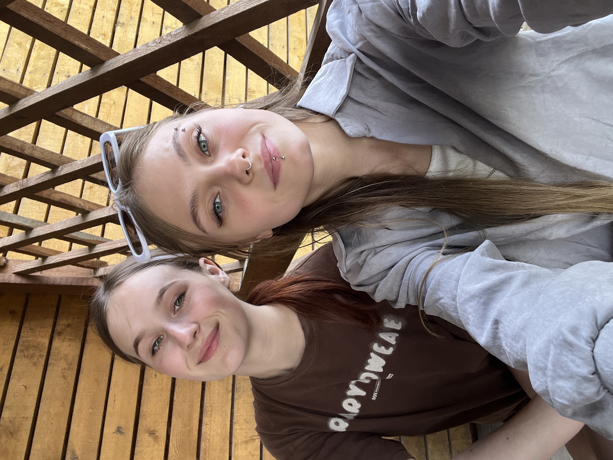 Two young women are posing for a selfie in what appears to be an outdoor setting, potentially a patio or deck, constructed from wooden planks. The woman on the upper portion of the frame has light brown hair, sunglasses pushed up on her head, and facial piercings (eyebrow, nose, lip). She wears a light-colored gray jacket. The other woman, positioned below, wears a brown shirt with the word "HAPPYWEAR" printed on it. Her hair is reddish-brown. The background consists of wooden planks and a lattice-like structure, suggesting an outdoor location. It is not possible to determine the precise location in the city of Фоки, Russia from this image.
