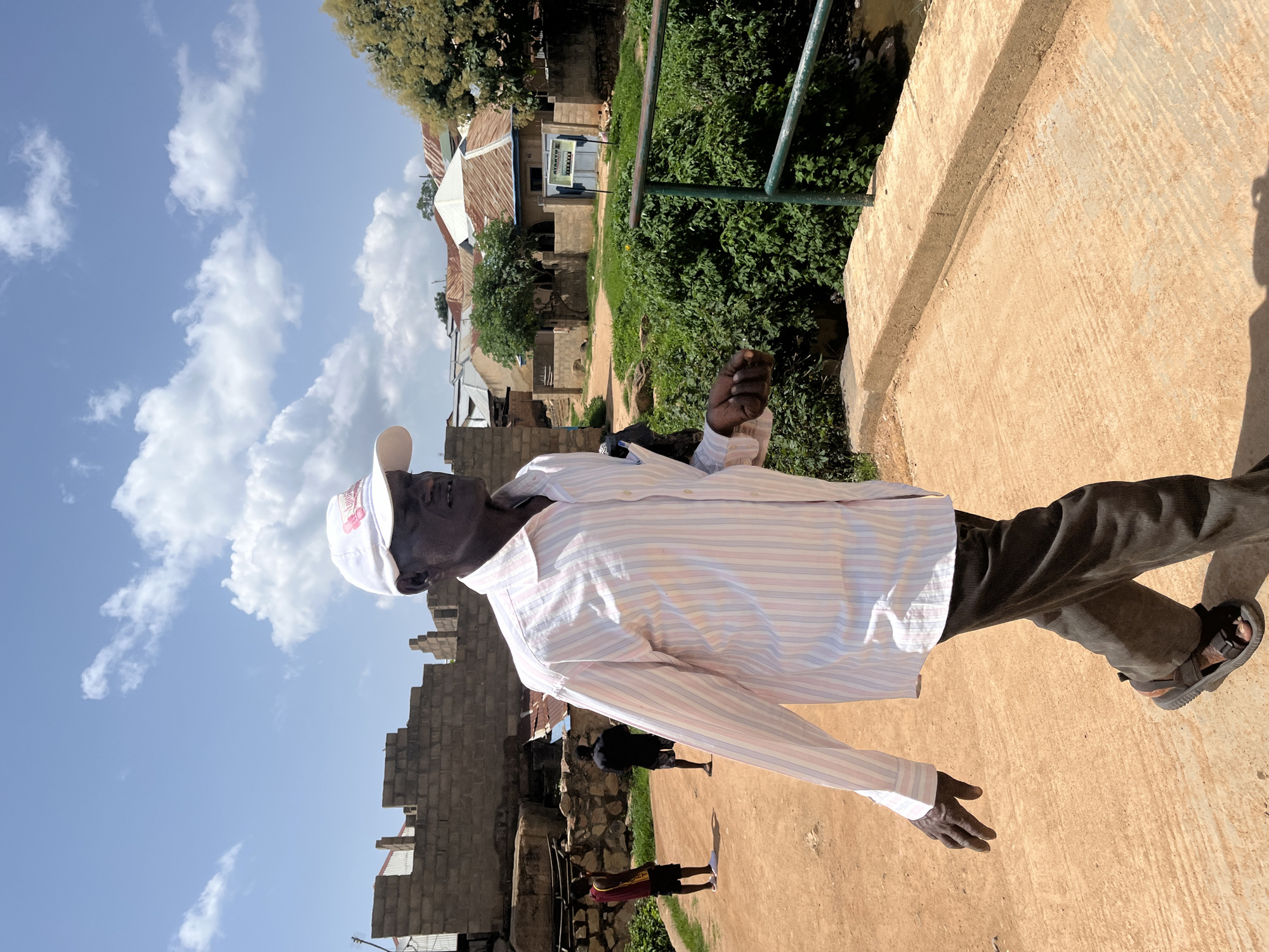 A man wearing a white baseball cap and a striped long-sleeved shirt walks on a concrete bridge in Jos, Nigeria. He is wearing dark brown corduroy pants and sandals. The man is looking to his right. In the background, there are unfinished brick buildings, trees, and houses with corrugated metal roofs. Two young men are visible in the distance on a dirt path. The sky is blue with scattered white clouds. A green metal railing runs along the side of the bridge.