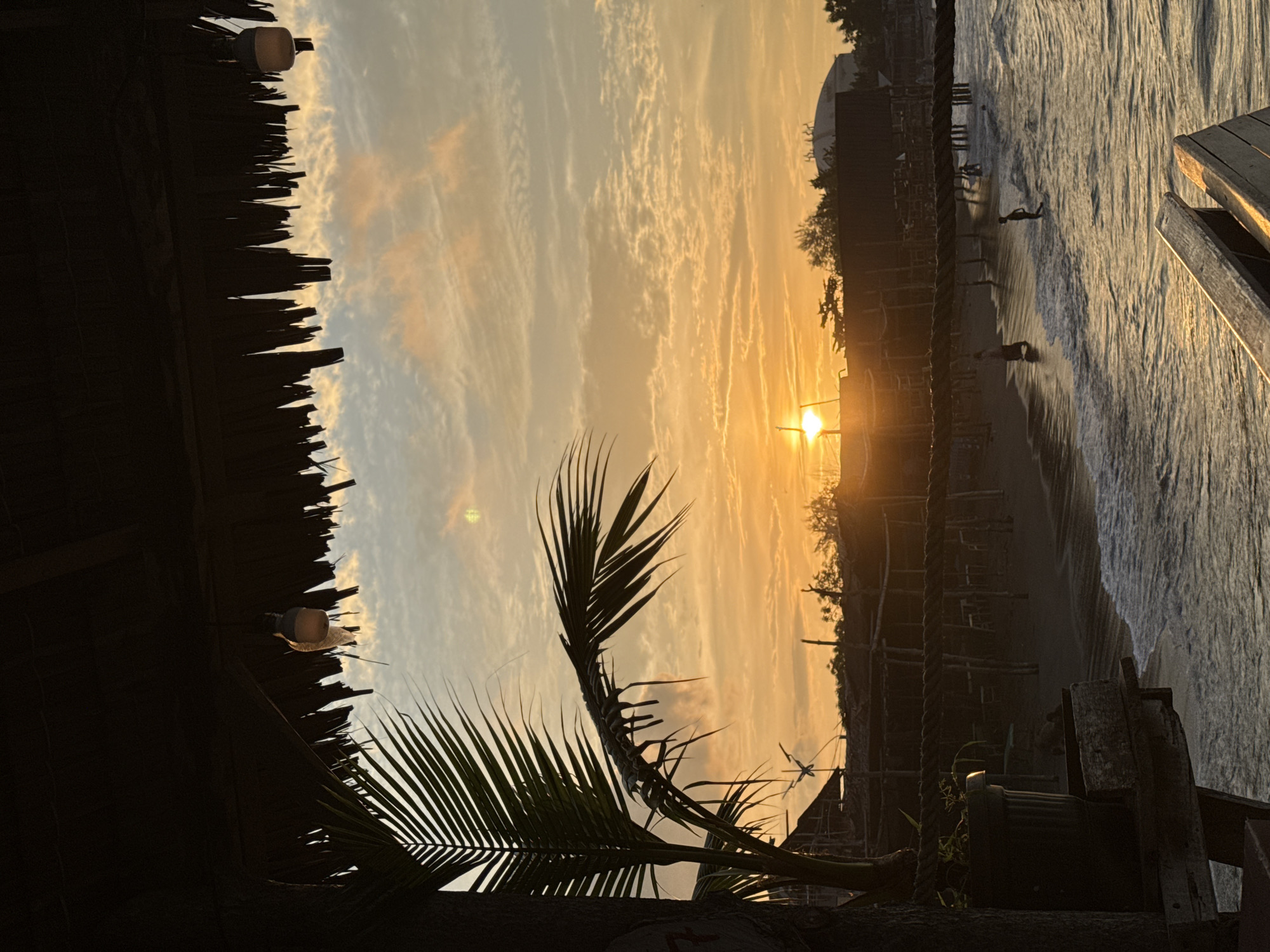 The image depicts a serene beach scene at sunset, likely in the city of Lhokseumawe, Indonesia. The sky is ablaze with warm hues of orange and yellow, characteristic of a tropical sunset, with scattered clouds catching the light. In the foreground, a thatched roof structure overhangs, with two simple hanging lights visible. To the left, the silhouette of a palm frond frames a portion of the view.
Below the structure, a sandy beach stretches along the coastline, with gentle waves lapping at the shore. Several small structures, possibly stalls or huts, are visible along the beach, supported by wooden posts. A few people are scattered along the water's edge and in the shallow sea; one individual walks along the wet sand, another stands in the water, and a third appears to be playing near the water.
In the mid-ground, a thick rope is strung horizontally, separating the immediate foreground from the beach area. A potted plant sits on a wooden surface in the lower left, partially obscuring the view of the beach. In the background, the silhouette of trees and distant buildings, including a dome-shaped structure, can be seen against the vibrant sky. The overall atmosphere is peaceful and tranquil, capturing the beauty of a coastal evening.