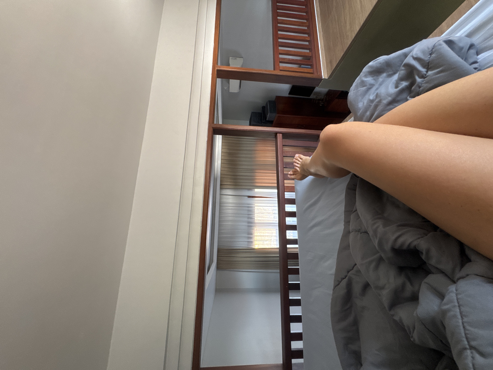 A first-person perspective shot from a bed shows a person's legs crossed and a grey comforter. Beyond the bed, wooden railings separate the sleeping area from a hallway. A window with sheer curtains is visible in the distance, with sunlight streaming through. To the right, a loft area with wooden railings and a wooden floor is visible. This scene is located in North Kuta, Indonesia.