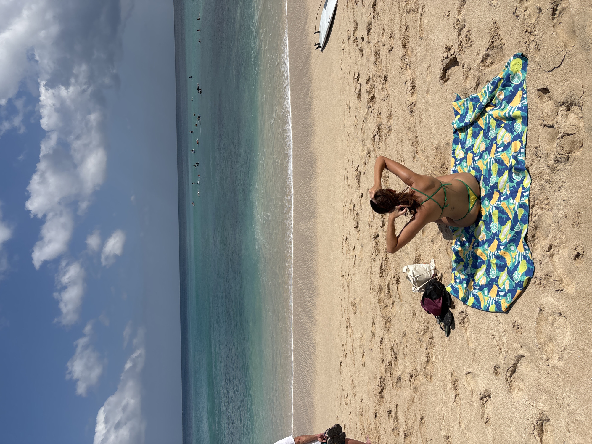 The image depicts a beach scene with several people visible. In the foreground, a woman is lying on a colorful beach towel with a fruit pattern. She is wearing a green bikini and has her hands behind her head. Next to her are a white bag and a dark backpack. A white surfboard is lying on the sand further up the beach. In the ocean, there are several surfers scattered across the water. The sand is a light brown, and there are footprints visible. The water is a clear turquoise color, with white waves breaking near the shore. The sky is blue with some scattered white clouds, suggesting a sunny day. The lighting indicates it is likely daytime. The scene appears to be a popular beach destination, possibly in a tropical location, given the clear water and the presence of surfers. The overall mood seems relaxed and typical of a day spent at the beach. There is no visible text in the image.