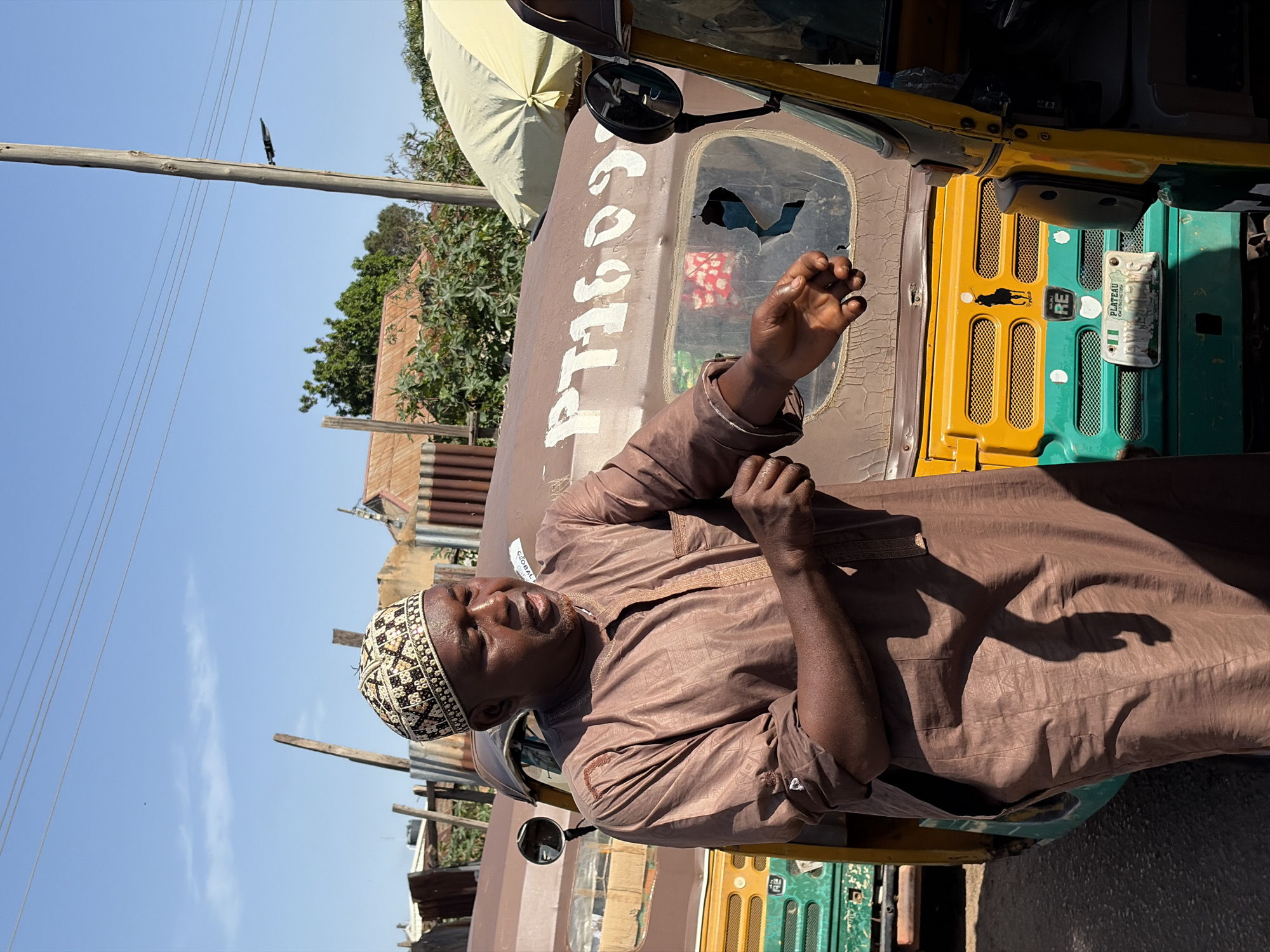 In the city of Gura Top, Nigeria, a man stands beside two auto-rickshaws under a clear blue sky. He is wearing a brown traditional outfit and a patterned cap, and appears to be speaking or gesturing. The auto-rickshaws are a mix of brown and yellow, with green accents. The one on the right has a broken window. In the background, there are various structures, including corrugated metal fences, a wooden pole with wires, and some vegetation. The overall scene suggests a street or a market area.