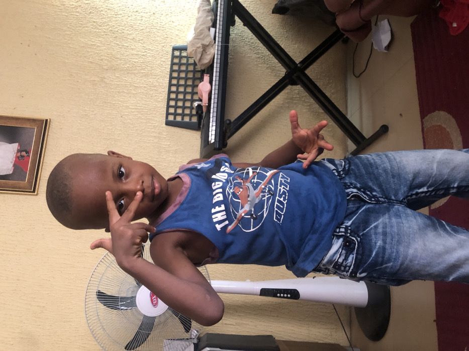 The image captures a young child standing indoors in Nkwelle Ezunaka, Nigeria. The child, of dark complexion and with short hair, is centrally positioned, looking directly at the viewer. The child's right hand is raised, with the index and middle fingers extended in a "V" shape near the face, while the left hand is held open to the side. The child wears a blue tank top adorned with a graphic of an airplane and the text "THE BIGGEST DUSTY," and faded blue denim jeans.
The background features a textured, light-yellow wall. To the child's left, a white standing fan with black blades and the "OX" logo is visible. To the child's right, an electronic keyboard rests on a black X-stand. Objects on the keyboard include a black grid-like component, a pink item, and light-colored crumpled material. Above the keyboard, a framed picture hangs on the wall, showing the lower portion of a person wearing white attire and red shoes. The floor consists of light-colored tiles, partially covered by a maroon rug with a circular pattern.