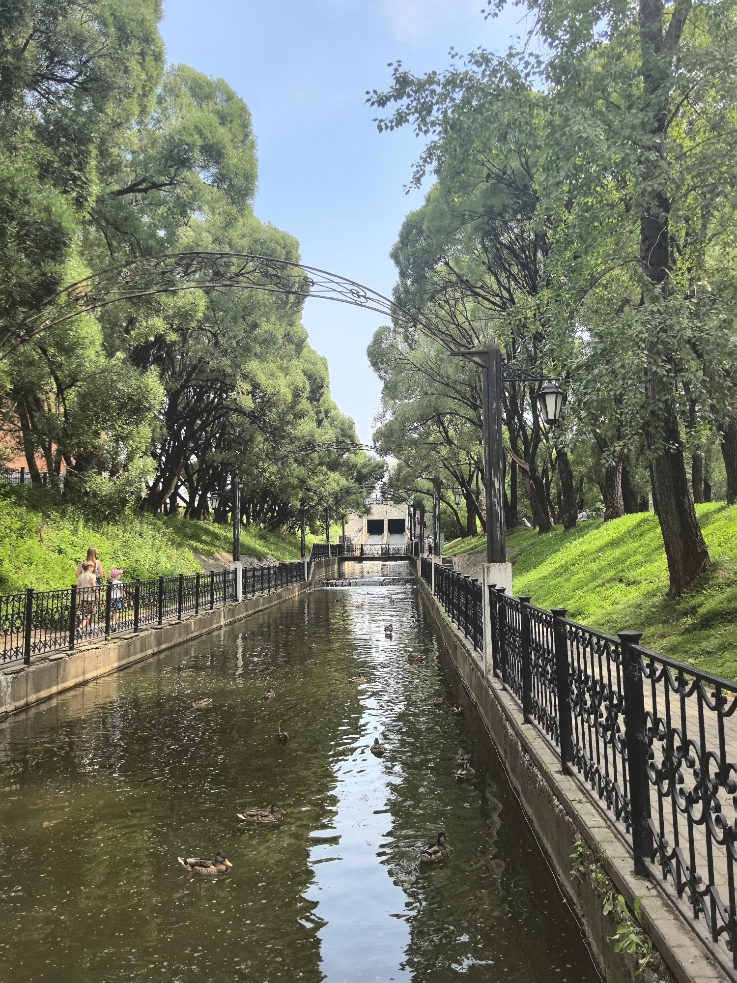 The image depicts a canal with ducks swimming in it, flanked by a pedestrian walkway and a grassy slope covered in trees. On the left side of the canal, there are two people walking on the path, a woman and a child. The walkway is bordered by a black metal fence with decorative elements. Street lamps are visible along the fence. The canal leads to a building with an arched structure in the background. The scene is set in a park-like environment with lush green vegetation and mature trees. The sky is clear and blue. This image was taken in Perm, Russia, at the embankment of the Kama River.