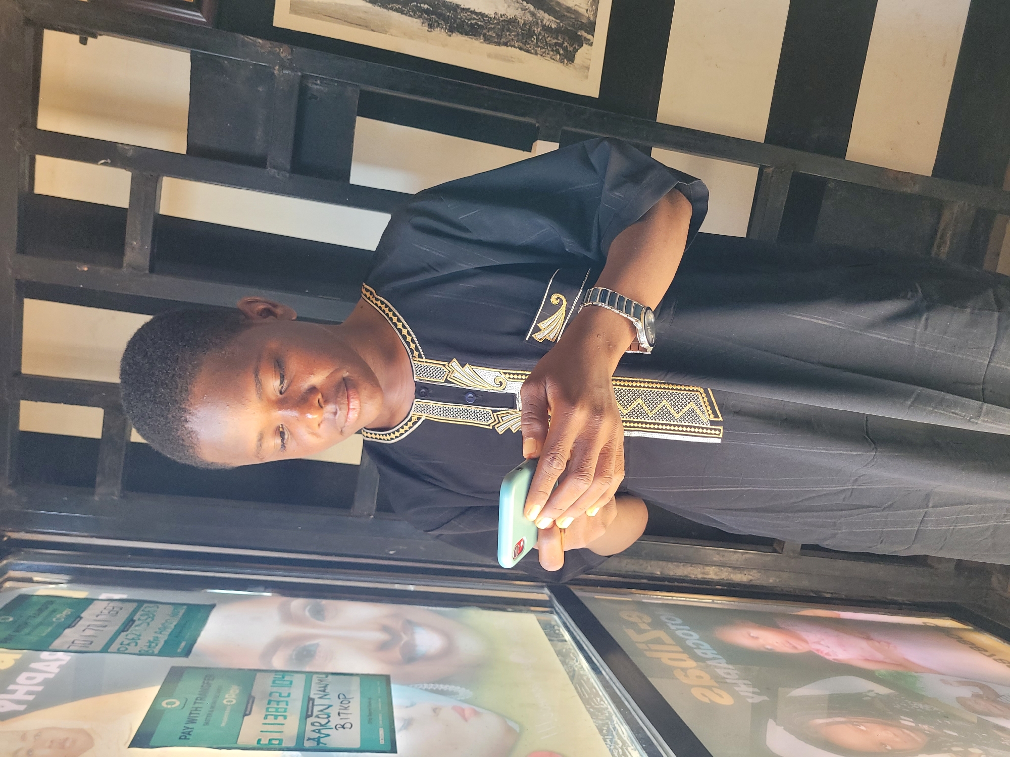 An upright, eye-level shot captures a young man indoors, likely in Jos, Nigeria, from the chest up. He has dark skin and short, dark hair, and is looking down intently at a light green smartphone held in his right hand. A silver-toned watch adorns his left wrist. He is dressed in a black tunic, featuring elaborate gold and black embroidery patterns around the neckline, on the upper chest, and on the sleeves, a style commonly seen in West Africa.
The background includes a dark, lattice-like partition or shelving unit, behind which a white wall and a framed picture are visible. In the foreground, below the man, is a reflective surface, likely a glass counter or window, displaying various advertisements and payment information.
Visible text on these displays indicates "PAY WITH TRANSFERS" options, suggesting a commercial transaction is taking place. One display shows "6113832104" for "AARON NAZILU BITKOP" via "Opay". Another offers "701 701 1667" and "P502500053" for "Opay", with the name "Fidelis Adom Nuhu" partially visible. The man appears focused on his phone, possibly facilitating a mobile payment or interacting with an application. The setting suggests an indoor commercial establishment during daytime.