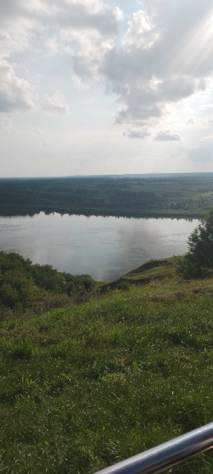 The image shows a wide river with a forest on the opposite bank. The foreground is a grassy slope, with a metal railing visible at the bottom right. The sky is partly cloudy, with sunlight breaking through. The scene is likely from a viewpoint overlooking the Belaya River in Birsk, Russia.