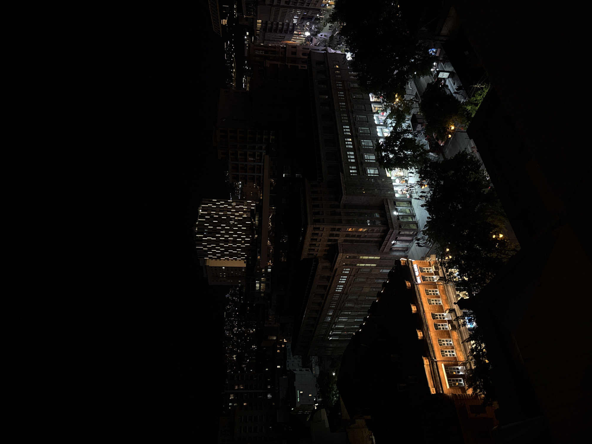 An eye-level, low-light, rotated photograph captures a cityscape at night. Buildings with illuminated windows are the main subject. A street scene with pedestrian activity is also visible. The buildings appear to be located in Yerevan, Armenia.