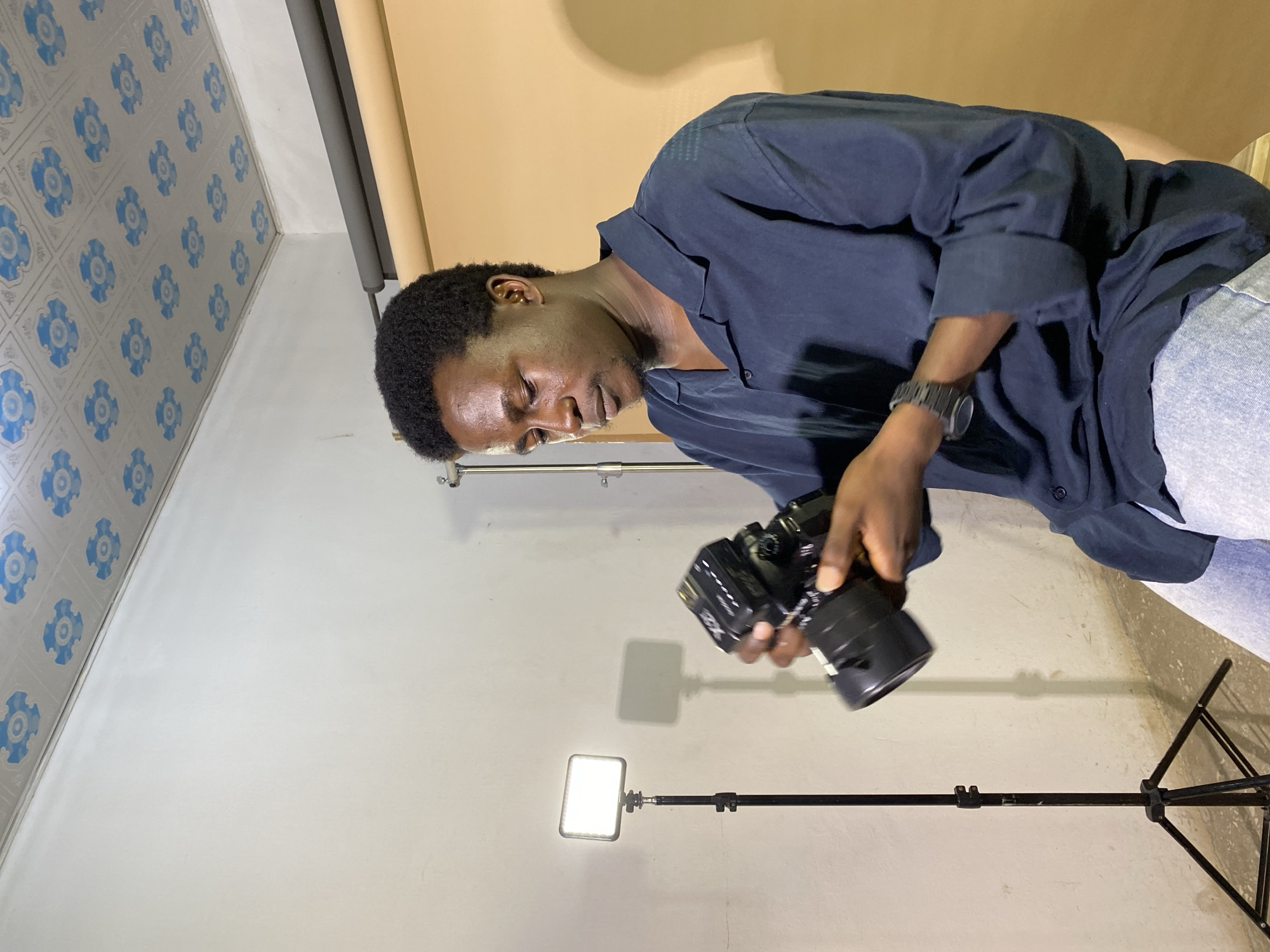 A young Black man, with dark, short, curly hair and a light beard, is seated in what appears to be a makeshift photography studio. He is wearing a dark blue, collared, button-down shirt and light blue jeans. He is holding a black DSLR camera with a lens attached, looking down at it as if to adjust settings or check something. On his left wrist, he wears a dark-colored watch.
To his left, a black tripod-mounted LED light panel is illuminated, casting a bright, square light onto the wall behind it, creating a distinct shadow. Further to the left, a section of a white wall is visible, with a ceiling above that features a repeating pattern of blue and white floral designs.
Behind the man, a backdrop made of a rolled-up tan or light brown fabric is visible, suggesting a controlled shooting environment. The floor is not clearly visible, but the overall impression is of an indoor space set up for photography, possibly in a residential area or a small business in Gura Top, Nigeria. The man appears focused on his camera, engrossed in his task.