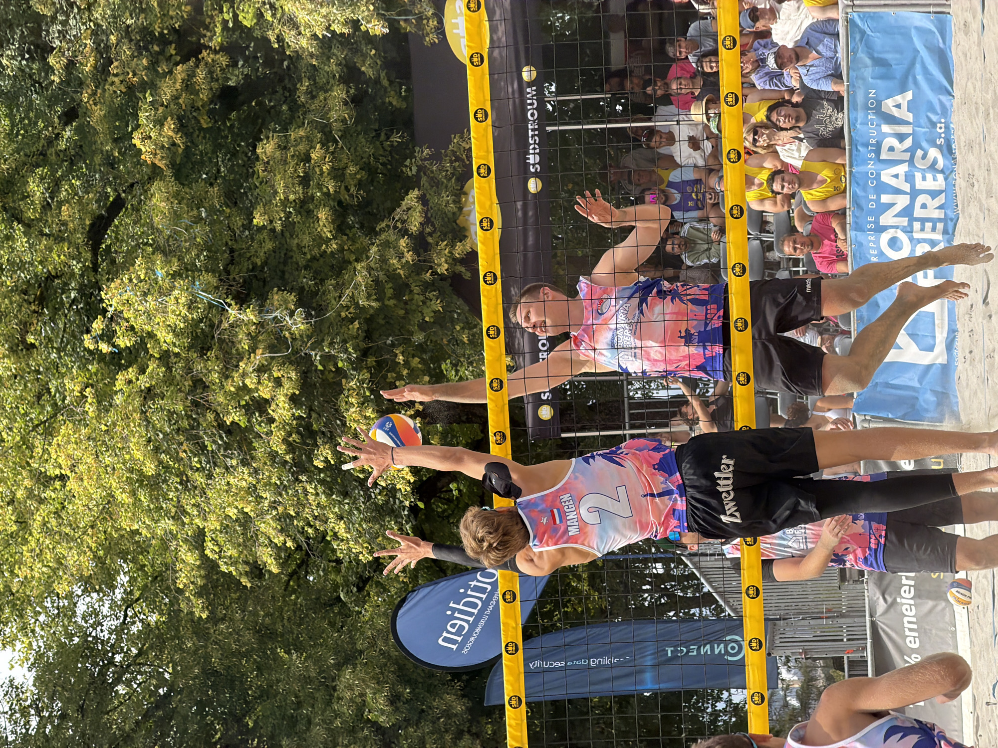 The image depicts a beach volleyball match in progress at the Bekaach Beach in Esch-sur-Alzette, Luxembourg. Two male athletes, wearing colorful tank tops with palm tree and cityscape graphics, are actively engaged in the game. The athlete on the left, wearing a tank top with the number "2" and the name "Mangen" on the back, is in the process of blocking a volleyball. The athlete on the right is also reaching for the ball. Both are positioned near a volleyball net, which is marked with yellow tape and logos. Spectators are visible in the background, seated behind the net. The environment consists of a sandy court with trees providing shade in the upper portion of the image. Sponsorship banners with text in French are displayed around the court, including "sudstroum" and "BONARIA FRERES s.a.". The overall scene captures a moment of athletic competition on a sunny day.