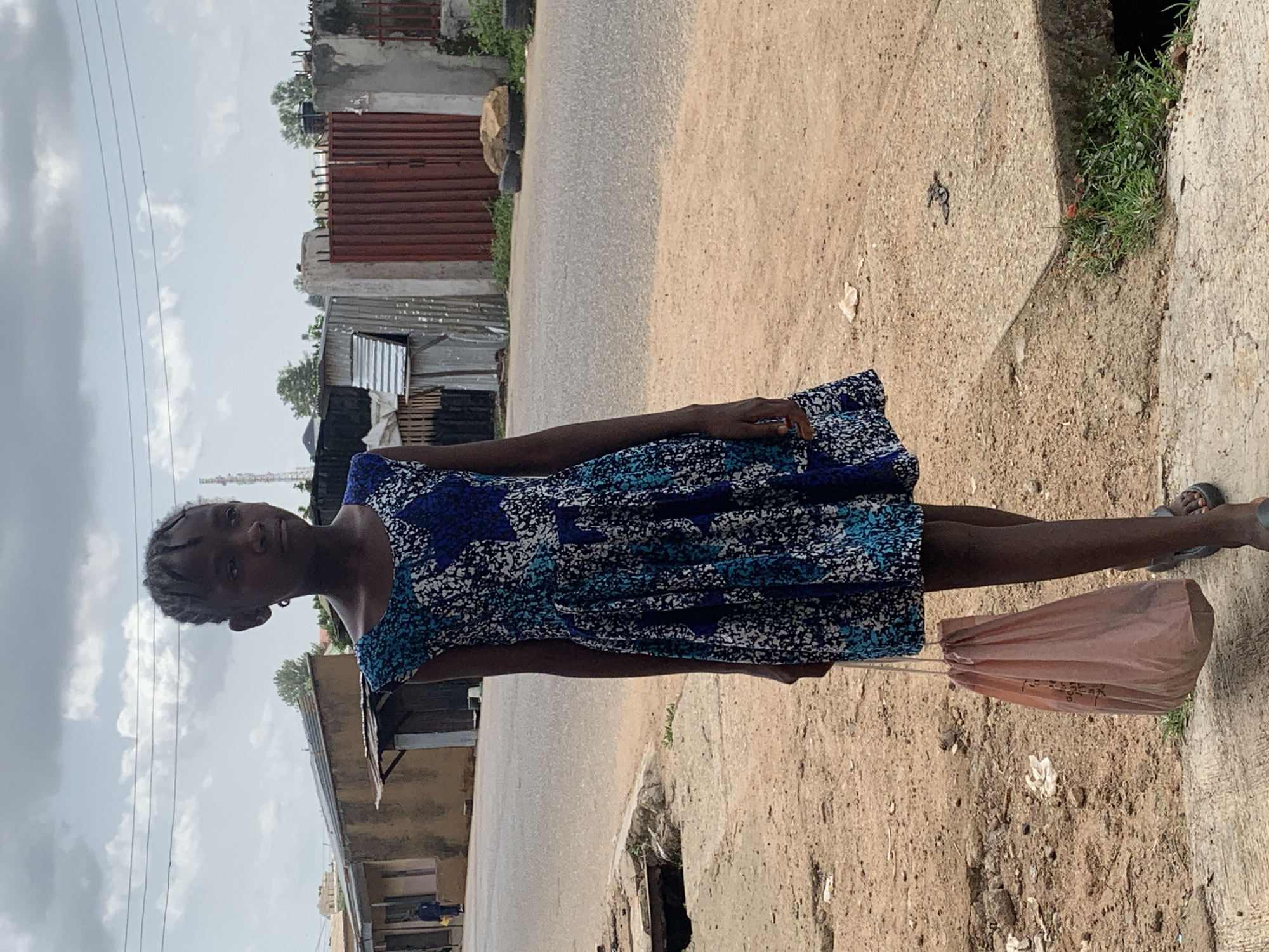 In Jos, Nigeria, a young Black girl stands on a dirt and concrete path next to a road. She is wearing a blue and white patterned dress and carrying a pink bag in her left hand. Her hair is braided and styled in small cornrows. In the background, there are buildings made of corrugated metal and concrete, some with red metal doors. A cell phone tower is visible in the distance. The sky is cloudy, with patches of blue peeking through. The ground is uneven and littered with debris.