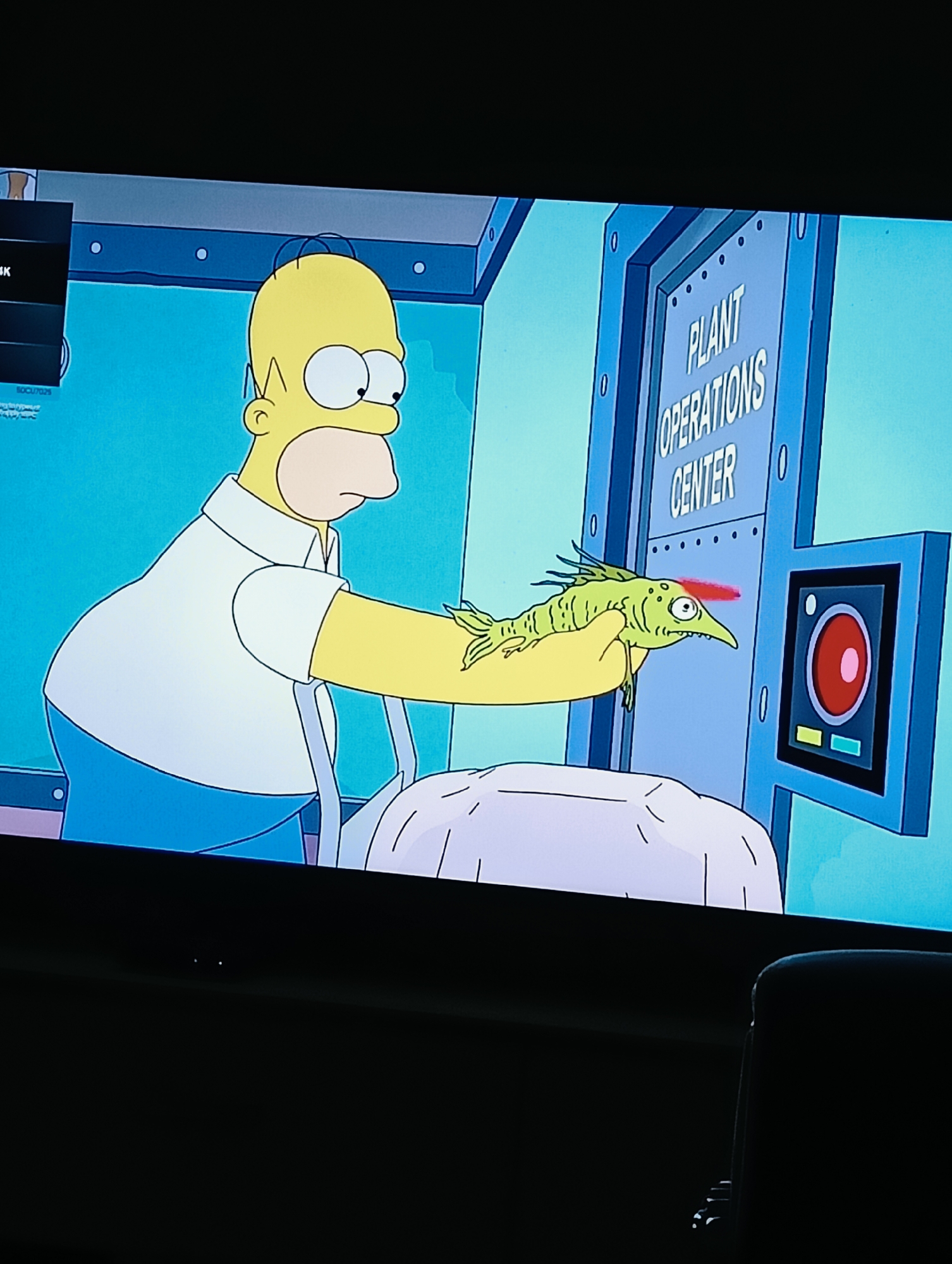 A television displays an episode of "The Simpsons" featuring Homer Simpson holding a three-eyed, green fish up to a scanner labeled "Plant Operations Center." The environment is a control room setting within the Springfield Nuclear Power Plant. The city of Dinkata, Bulgaria is not visible in this image.