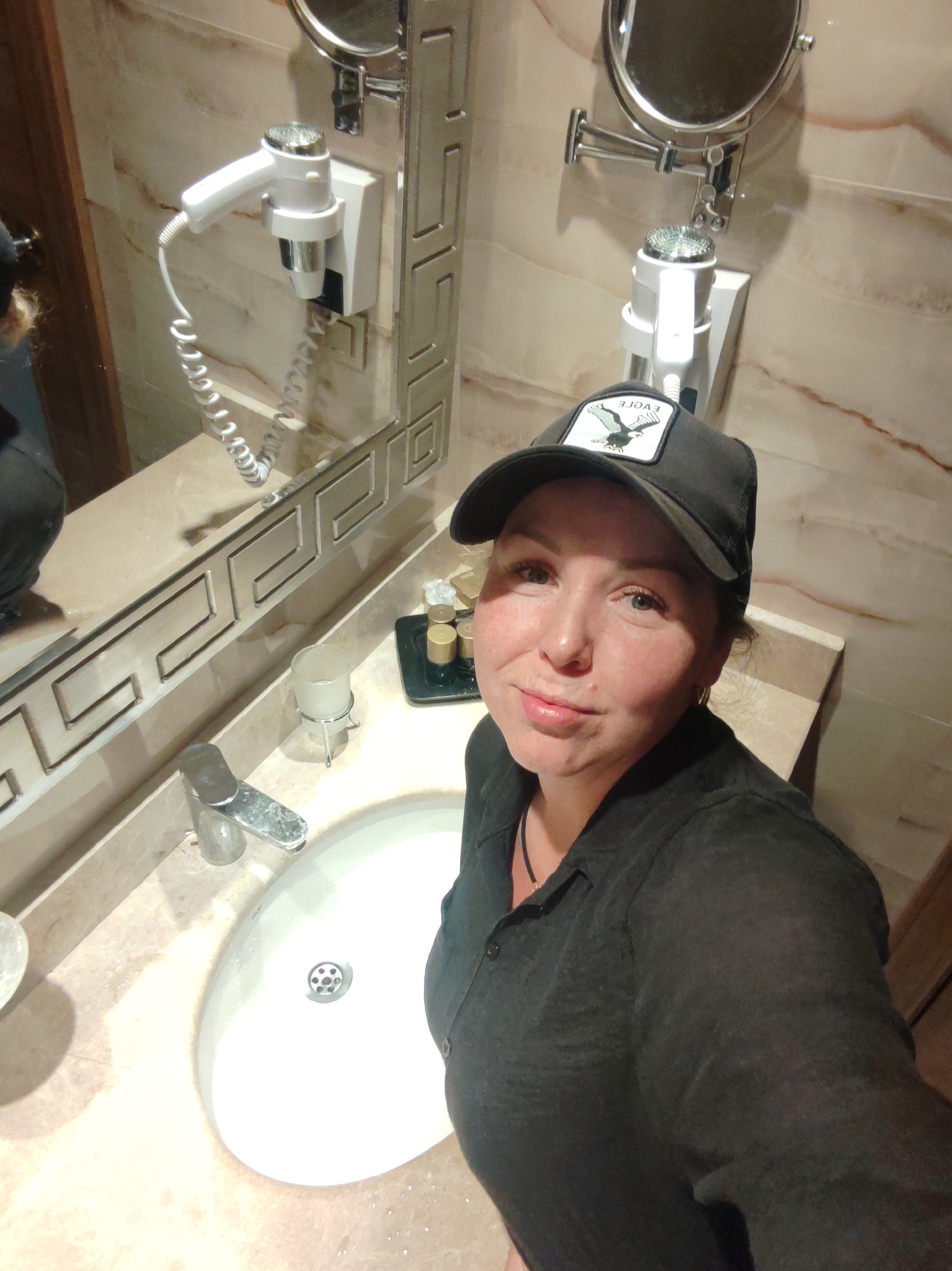 A woman wearing a black baseball cap and a dark-colored shirt is taking a selfie in a bathroom. The bathroom has a marble countertop with a white sink, chrome faucet, and toiletries. There's a large mirror with a decorative frame above the sink and a wall-mounted hairdryer and magnifying mirror on the adjacent wall. The location is Unknown, Unknown.