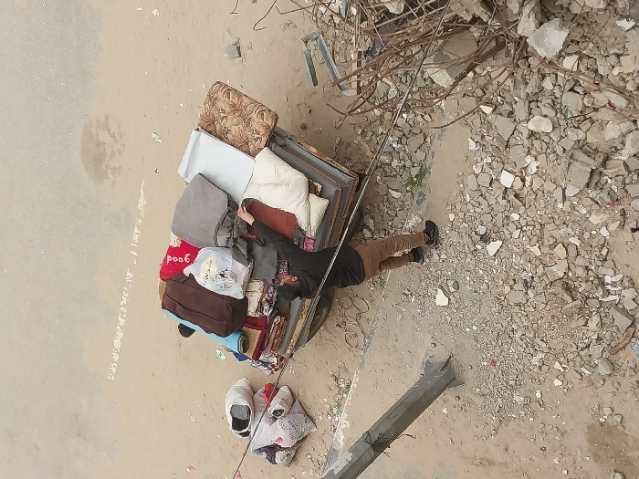 An overhead view captures an adult male interacting with a heavily laden utility vehicle or cart in Khan Yunis, Palestinian Territory. The male, dressed in a dark long-sleeved top, light brown trousers, and dark shoes, stands to the left of the vehicle's bed, reaching with his right hand to adjust or secure items. The vehicle bed is stacked with household goods, including a red blanket with "good" visible text, a gray blanket, a white patterned blanket, a dark brown bag, a plain white cushion, a floral-patterned cushion, a metallic panel, and several plastic bags, one partially displaying Arabic text. To the left of the vehicle, additional bundled items in white bags rest on the ground. The surrounding environment consists of unpaved, light brown earth extensively mixed with concrete rubble and scattered debris. A light-colored utility pole or beam is visible on the left side of the frame, embedded in the debris. To the right, a substantial pile of twisted rebar and other structural wreckage indicates a destroyed area. The background shows a broad expanse of light-colored, unpaved ground, likely a dirt road or open area.