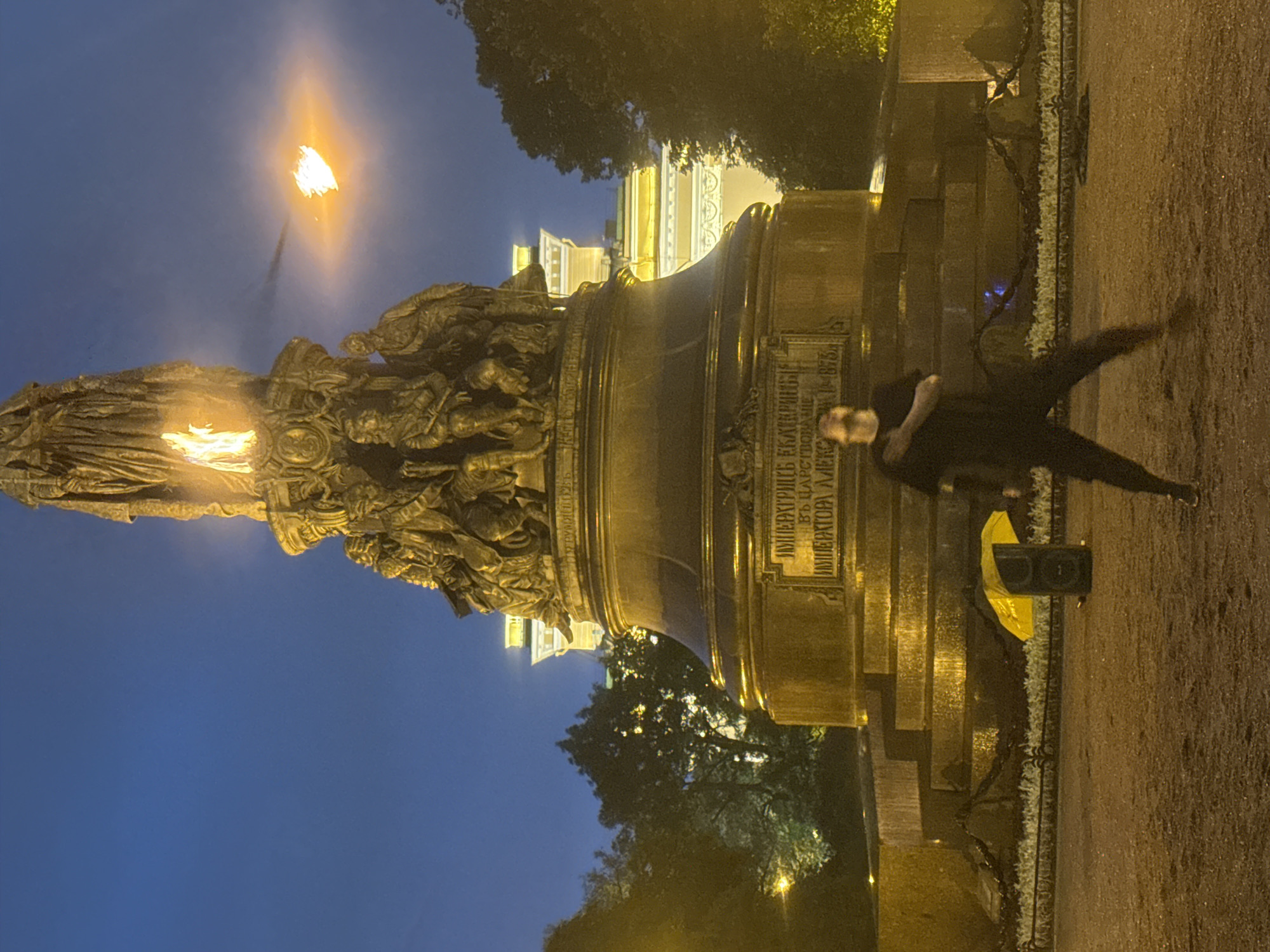 A person in dark clothing, blurred by motion, is captured in the foreground of the image. They are positioned in front of a large, ornate bronze monument, which is illuminated by flames at its top and on a torch extending from its side. The monument is detailed with sculpted figures and inscriptions. Behind the monument, a building with glowing yellow lights is visible. The scene is set at dusk or night, with a deep blue sky. The ground is a dirt or gravel surface. To the left of the monument, a dark speaker box sits on the ground, partially obscured by a bright yellow umbrella. The environment appears to be an outdoor public space, likely a park or square in Saint Petersburg, Russia, given the architectural style and the monument's appearance, which strongly resembles the Monument to Catherine the Great in Nevsky Prospekt.