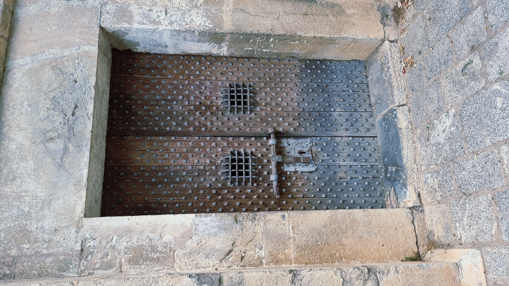 The image shows a heavy, weathered wooden double door set within a stone archway. The doors are studded with numerous small, square metal rivets. Each door has a small, square metal grate set into its upper half. A thick, horizontal metal bar slides through two latches mounted on the doors, securing them. One latch has a keyhole visible. The stone surrounding the door appears aged and textured, with some evidence of wear and chipping. The ground in front of the door is paved with rough cobblestones. The setting appears to be an exterior entryway. The location is the city of Montagut i Oix, Spain, specifically at the Church of Sant Pere, where this imposing door is a notable feature. There are no people or animals visible in the image.