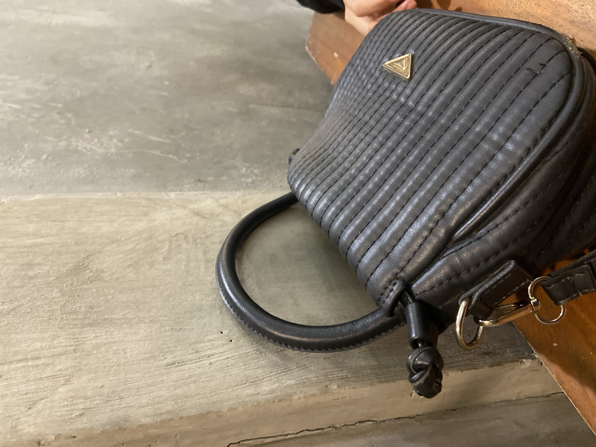 A black quilted handbag with a triangular metal emblem rests on a wooden surface against a textured concrete wall. The bag has a single looped handle and a detachable strap attached with metal clips. The background is dimly lit, with the concrete wall showing slight variations in tone and texture. A portion of a person's hand is visible on the right side of the image, suggesting the bag may belong to them. The overall scene appears to be indoors, potentially in a cafe or public space. The image does not provide any information about the city of Sekar Mawar, Indonesia.
