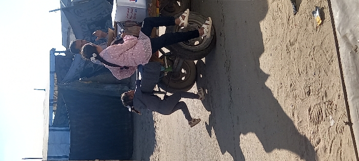 An outdoor scene in Khan Yunis, Palestinian Territory, depicts three individuals interacting with a two-wheeled cart on a dirt and sand surface under a clear sky. A young girl in a gray tracksuit and sandals is positioned on the left, actively pushing the cart from behind. Another girl, wearing a pink patterned long-sleeved top, dark pants, and white sandals, sits sideways on the cart's frame, with her legs dangling over one of the wheels. An adult male, partially visible behind the second girl, is also on or near the cart, appearing to hold something in his hand. The cart carries a white box with red and blue markings and Arabic text. In the background, several makeshift structures, constructed from tarpaulins and other materials, are visible, suggesting a temporary settlement. Long shadows are cast on the ground from the subjects and the cart, indicating direct sunlight.