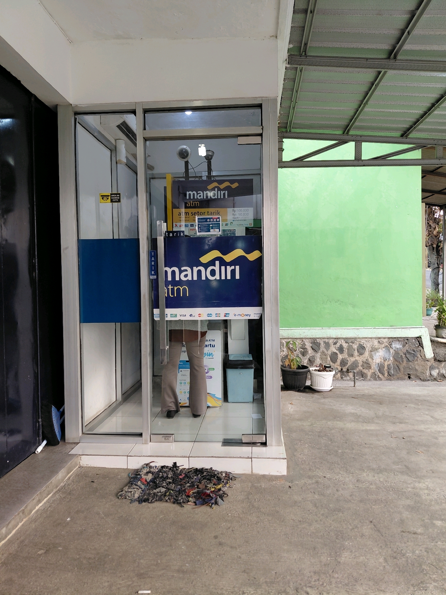 The image shows the exterior of a Bank Mandiri ATM booth located in Karanggede, Indonesia. The ATM booth has glass doors and walls, with a yellow and blue sign for Bank Mandiri visible on the ATM machine inside. A person's legs and lower torso are visible through the glass doors as they stand inside the booth. The surrounding area is paved with concrete, and there is a small pile of colorful rags on the ground in front of the ATM. To the right of the ATM, there is a light green wall, a stone-lined planter with plants, and a metal awning overhead. The scene appears to be outdoors during the daytime, with natural light illuminating the area.