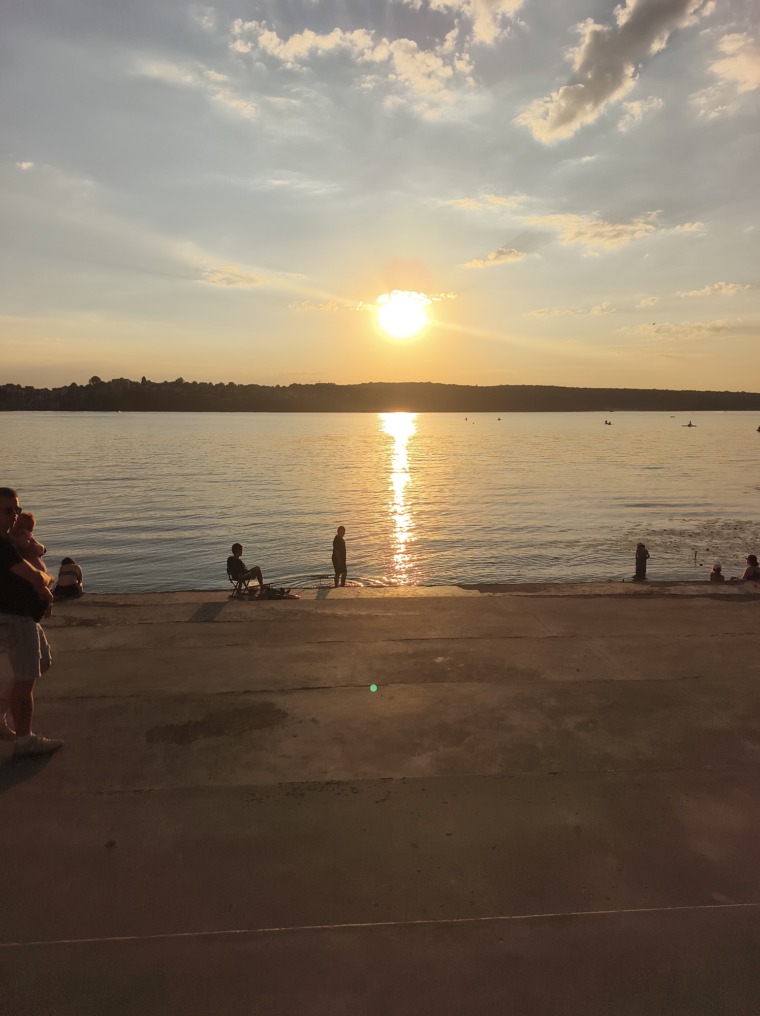 This image depicts a sunset over a body of water, likely Ternopil Lake in Ternopil, Ukraine. The sun is low in the sky, casting a warm golden glow across the water and illuminating a line of trees and hills on the far shore. Several people are visible along the water's edge, some sitting and others standing. In the foreground, a man with a baby on his hip is walking away from the camera. The water is calm, with a reflection of the sun creating a shimmering path. The overall scene is serene and evokes a sense of peaceful evening activity.