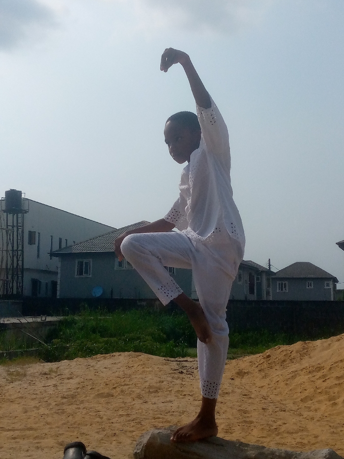 A person balances on one foot, poised on a weathered log amidst a sandy landscape. Dressed in white attire with eyelet detailing, the individual extends one arm upward, fingers curved, while the opposite leg is bent at the knee, mirroring the arm's position. Behind them, a green expanse separates the scene from nearby buildings, including a water tower, under a hazy sky.