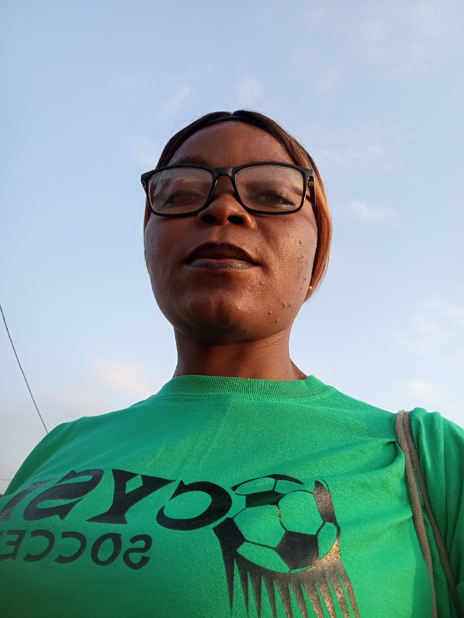 A low-angle, full-frame photograph captures a woman with dark skin, wearing glasses and a green t-shirt with a black graphic of a soccer ball and the word "SOCCER" in black lettering. The graphic is partially obscured by the woman's shoulder. She has dark hair that appears to be pulled back, with some strands framing her face and a reddish-brown highlight visible on the right side. Her makeup includes dark lipstick and eyeliner. She is looking directly at the camera. The background consists of a pale blue sky with some wispy white clouds, suggesting an outdoor setting during the day. A thin, dark electrical wire or string is visible on the left side of the frame, running vertically. A thin, light brown strap, possibly from a bag, crosses over her right shoulder and down her back. The image was taken in Kason, Nigeria.