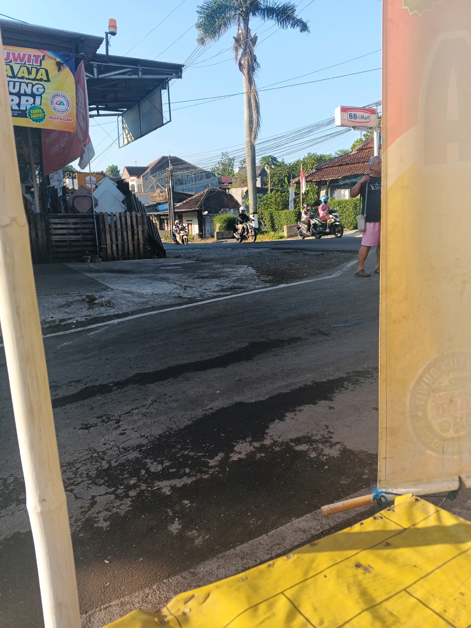 The image depicts a street scene in Semarang, Indonesia, likely during the daytime given the sunlight. In the foreground, a pale yellow, cylindrical object, possibly a bamboo pole, is positioned vertically on the left side of the frame, partially obscuring the view. A bright yellow tarp is visible in the bottom right corner, secured by a thin wooden dowel and blue rope.
The street itself is paved with asphalt, showing signs of wear and tear with patches of moisture. A white line marks the edge of the road. In the mid-ground, several motorcycles are present. One motorcycle, ridden by two individuals, is moving away from the camera. Another motorcycle, also with two riders, is stationary on the road. Further back, another motorcycle is seen from the side.
On the left side of the street, a covered stall or structure is visible with a yellow banner displaying Indonesian text and some logos. Wooden pallets are stacked nearby. A tall palm tree with a slender trunk dominates the upper center of the image.
On the right side of the street, a sign for "88 Mart," a convenience store, is mounted on a pole. A man wearing a dark t-shirt and pink shorts is standing near the sign, facing away from the camera. Behind the man, a building with a reddish-brown tiled roof is visible, surrounded by greenery. Electrical wires are strung across the sky. The overall impression is that of a busy local street in a tropical urban environment.