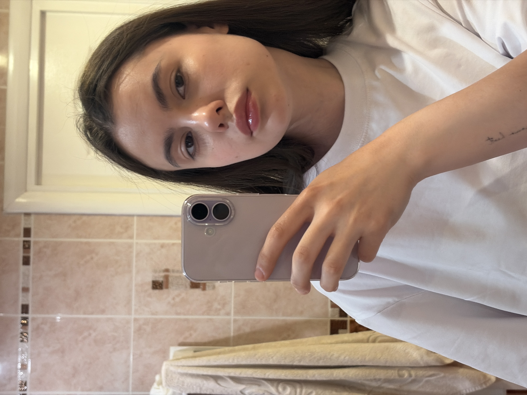 The image shows a young woman holding a smartphone in her right hand, taking a selfie in what appears to be a bathroom. She has dark hair and is wearing a white t-shirt. The background reveals beige tiled walls and a mirror, suggesting a bathroom setting. There are towels in the bottom of the image. The image does not provide information regarding the location in the city of Chaikovsky, Russia.