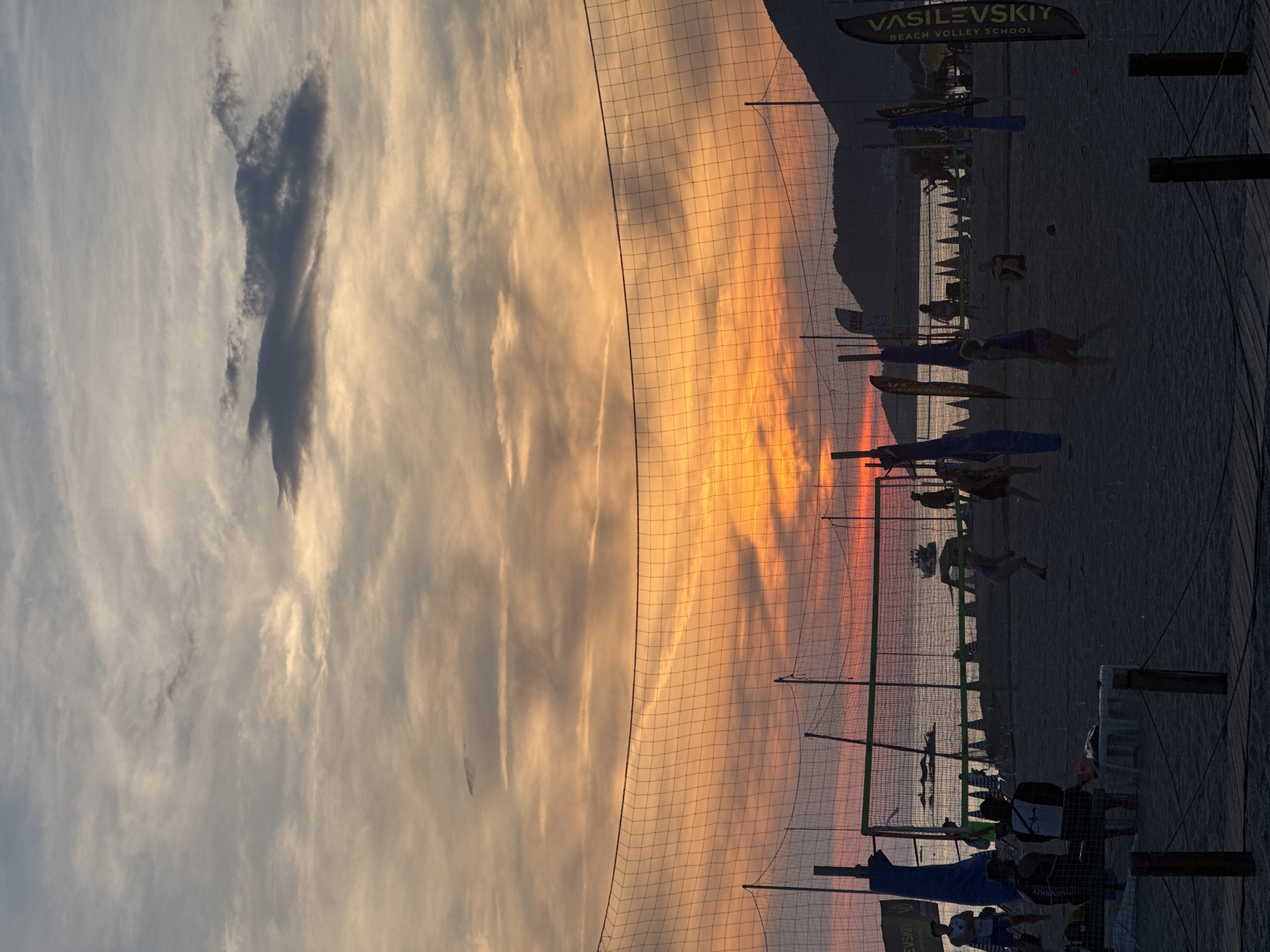 The image shows a beach scene at sunset. A volleyball net runs vertically across the center of the frame. The sky is a blend of orange, pink, and blue, indicating sunset. On the right side of the net, silhouetted figures are visible on the beach, with flags and what appears to be a beach volleyball school sign labeled "VASILEVSKIY BEACH VOLLEY SCHOOL." The beach appears sandy, and there's a slight indication of a hill or mountain range in the background. People are engaged in beach activities such as playing volleyball. The location appears to be near a body of water.