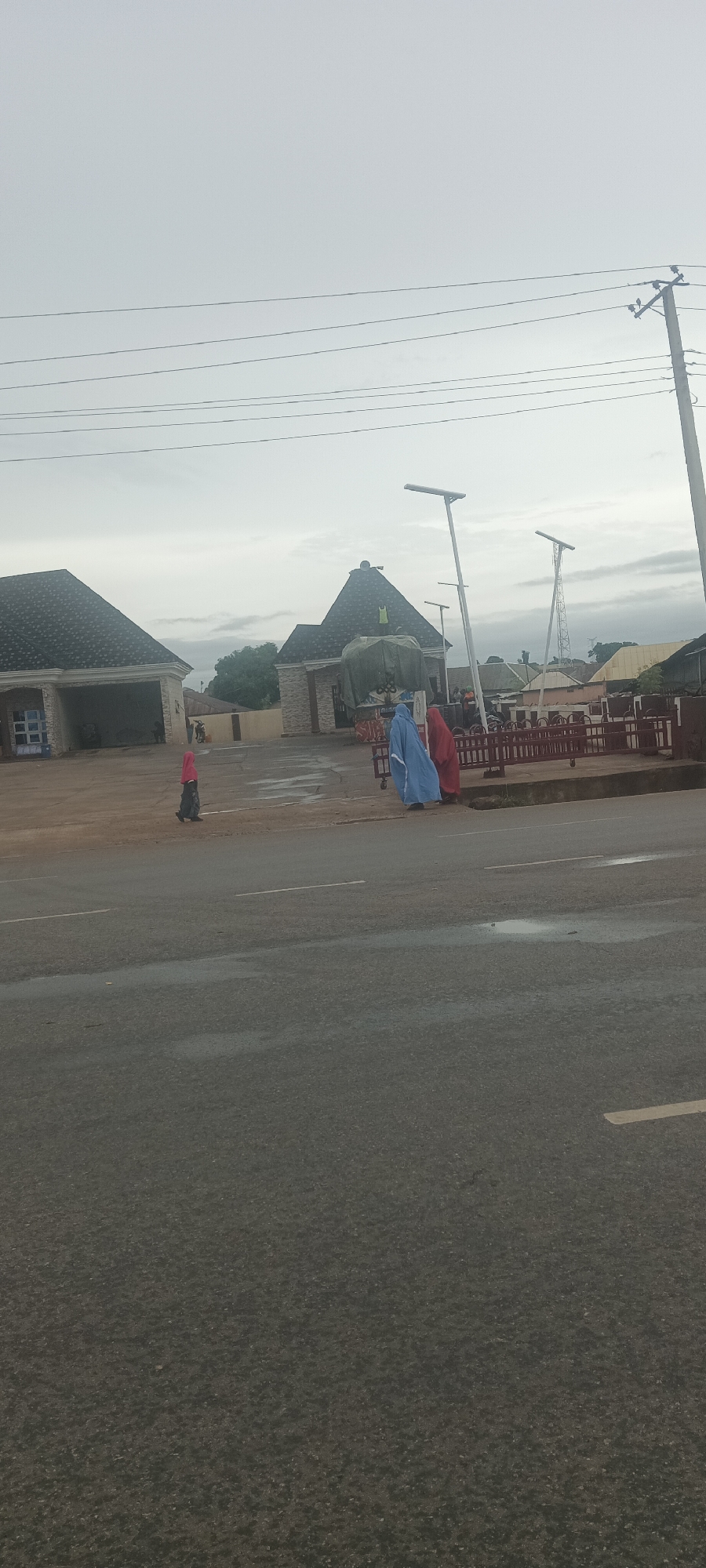 The image depicts a scene in Lafia, Nigeria, likely near a building with a gated entrance and what seems to be a cart selling items. Two people draped in blue and red garments are near the gate. Another person, wearing a red head covering, walks in the vicinity. Modern streetlights are in place. Power lines are visible above. The asphalt road in the foreground shows signs of minor water puddles. The sky is overcast.