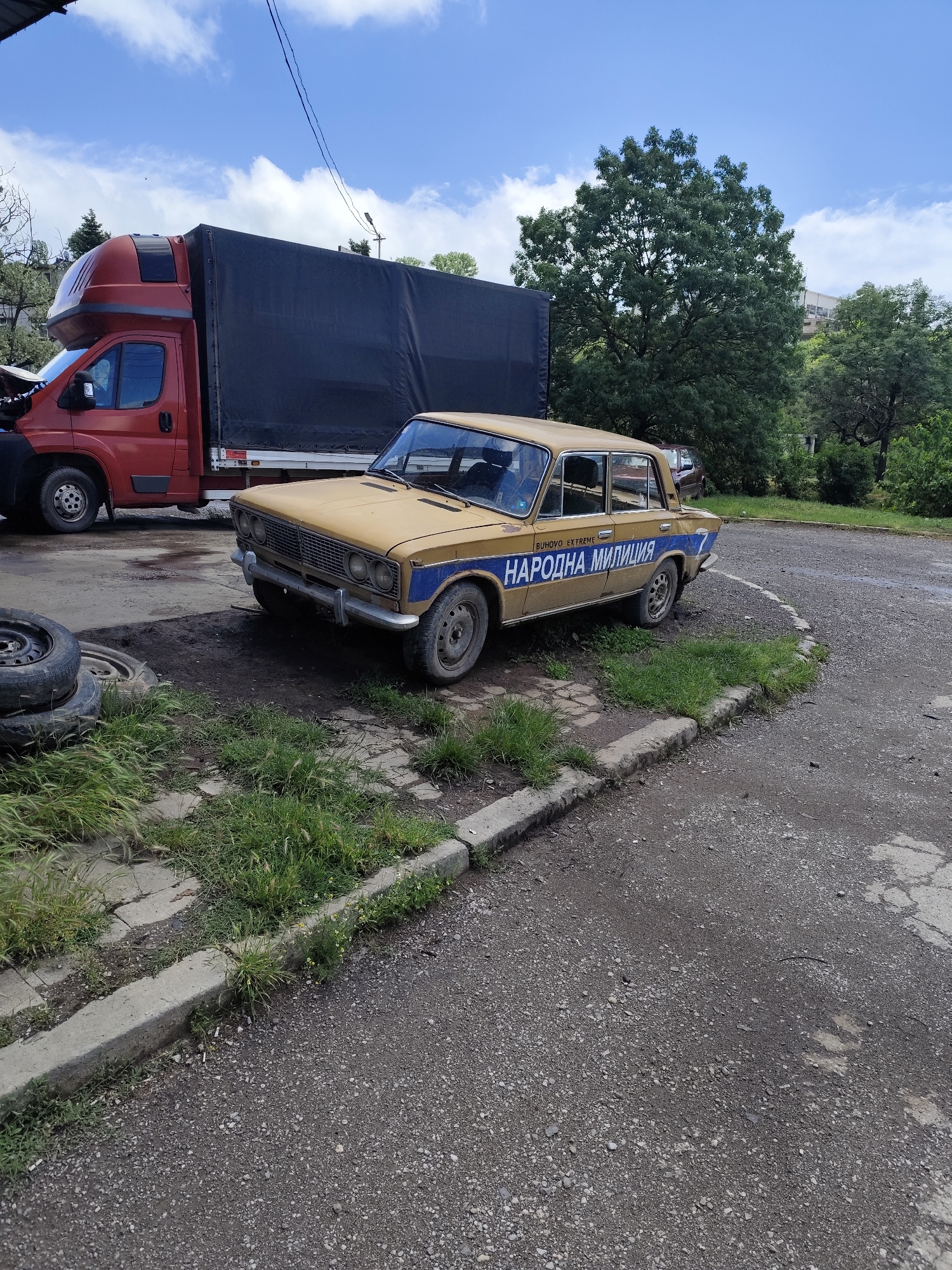 An outdoor, eye-level shot captures a static scene featuring two vehicles and surrounding terrain under a partly cloudy sky. The main subject is an old, weathered yellow four-door sedan, resembling a Lada, parked on a patch of dirt and sparse grass, partially overlapping a gravel path. A prominent blue stripe runs along its side, featuring text in Cyrillic. Above the stripe, "BUHOVO EXTREME" is visible, and on the stripe itself, "НАРОДНА МИЛИЦИЯ" (Narodna Militsiya, meaning People's Militia) is clearly written, followed by the number "7" towards the rear of the car. The car's paint is faded, and its wheels appear worn.
To the left and behind the yellow sedan is a large red box truck, possibly a Fiat Ducato model, with a black tarpaulin covering its cargo area. Its front hood appears to be open, suggesting it might be undergoing maintenance or repair. In the immediate foreground, to the left, two old, black tires are stacked on the ground.
The ground consists of a mix of gravel and dirt paths, separated by concrete curbs, with areas of overgrown green grass and weeds. In the background, numerous lush green trees rise towards a blue sky dotted with scattered white clouds, indicating daytime with fair weather. No people are visible in the image. The scene suggests an outdoor vehicle storage or repair area in Buhovo, Bulgaria.