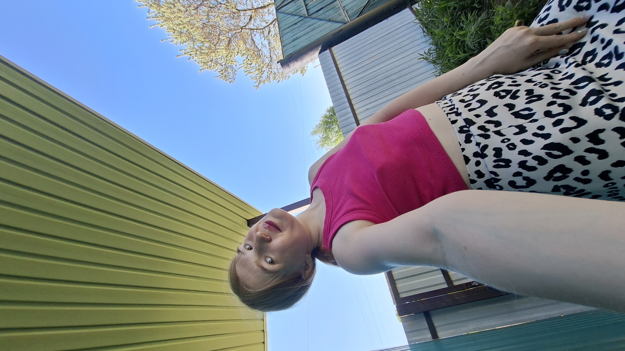 A low-angle shot shows a person in a pink top and animal-print pants lying on what appears to be a pool cover. The individual is fair-skinned with light hair and is looking towards the camera. The background includes a green corrugated metal wall on the left, a section of a blue sky, and foliage with tree branches at the top. The scene appears to be outdoors, with sections of buildings visible overhead. The picture was taken in daylight. While Большой Букор, Russia is mentioned, there is no specific detail within the image to confirm the location.