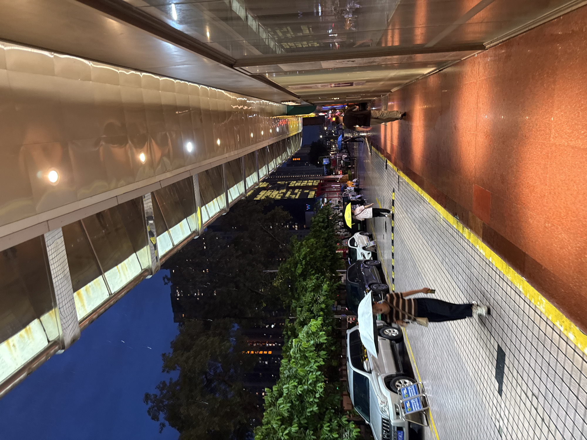 This image depicts a street scene in Tianhe District, China, at dusk or night. A covered walkway with reflective panels and overhead lighting extends along the right side of the frame, casting a warm orange glow on the wet pavement below. To the left, a sidewalk with a checkered pattern is bordered by a yellow stripe and leads to a street with parked cars. Beyond the street, lush green trees and tall buildings with illuminated windows are visible against the dark blue sky. Several people are walking under umbrellas, indicating it is raining. One person, a woman with dark hair, is walking towards the viewer under a light-colored umbrella, wearing a striped shirt and dark pants. Another person, a man with dark hair, is walking away from the viewer further down the covered walkway, wearing a dark t-shirt and light-colored pants. Other individuals and vehicles are present in the background, contributing to the bustling urban atmosphere.