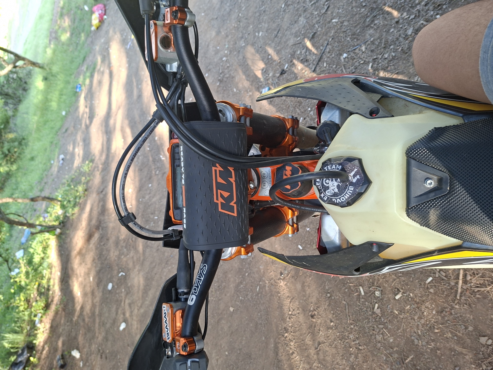 The image shows the top of a KTM 6 Days motorcycle, including the handlebars, front fender, gas tank, and the rider's leg. The motorcycle's handlebars have black grips with "6 DAYS" in white lettering on the left side and a black KTM-branded pad in the center. Orange handlebar clamps secure the handlebars. The gas tank is light yellow and has a black sticker with the text "TEAM BUHOVO EX." written on it. The seat has a black patterned cover. The motorcycle is situated on dirt ground with some scattered debris. The background is blurry and includes trees and grass, suggesting an outdoor setting. The leg of the person riding is on the top right of the frame. The OCR information indicates that the picture was taken in Buhovo, Bulgaria.