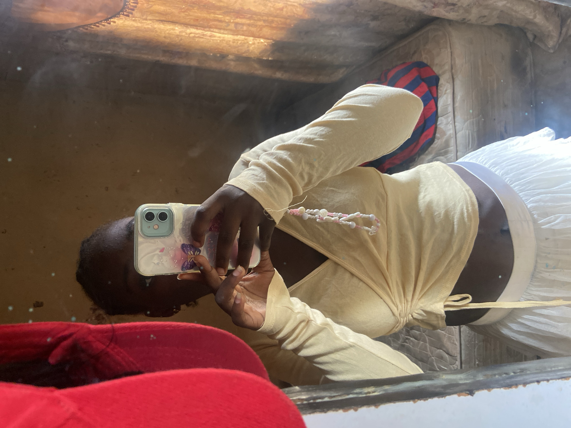 A person is taking a selfie in a mirror. They are holding a phone with a butterfly case. They are wearing a light yellow long-sleeved top and a white skirt. In the background, a mattress with a red and blue striped blanket is visible. The scene appears to be indoors, possibly in a bedroom. The lighting suggests it might be daytime.
