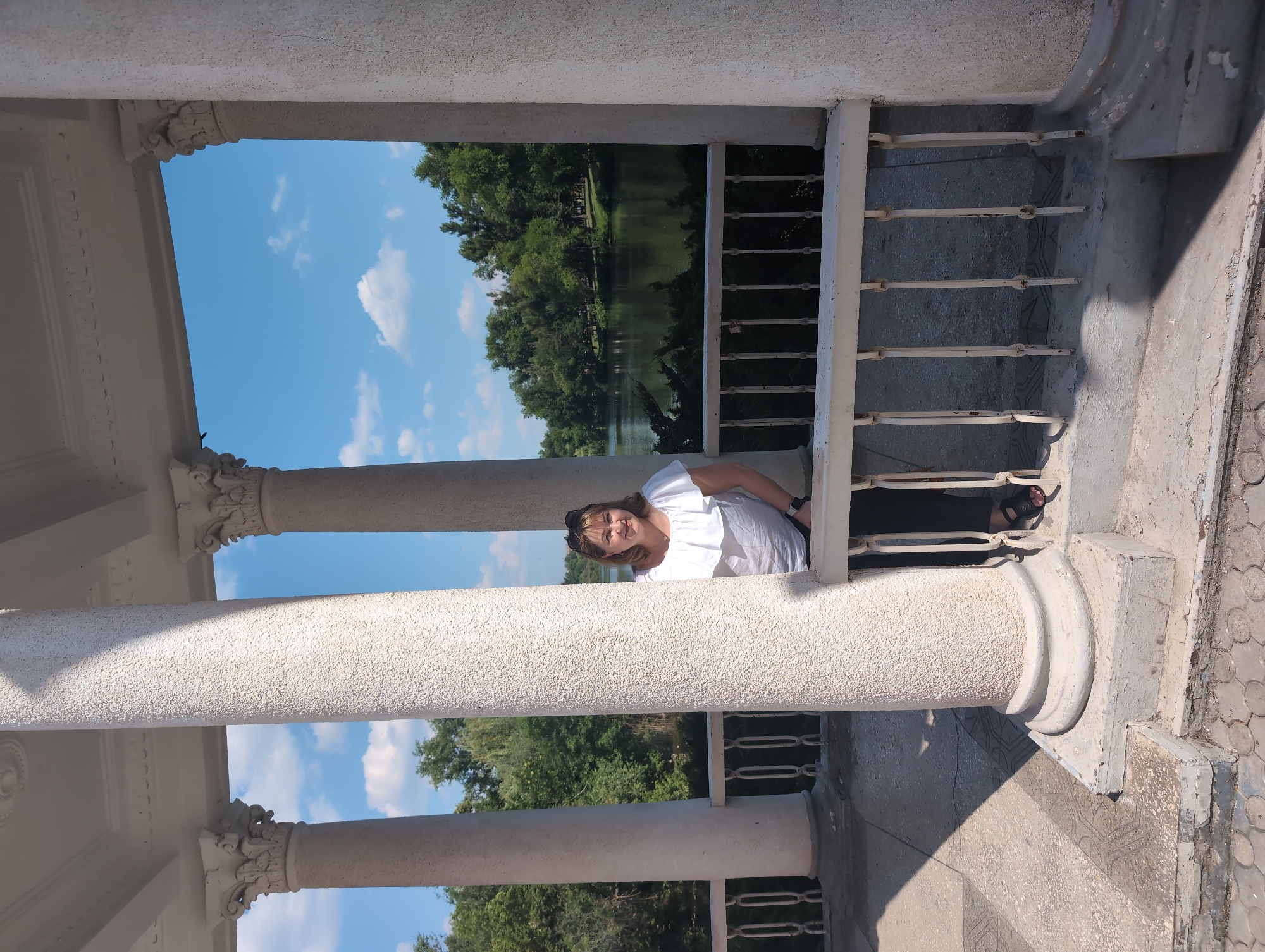 A woman is leaning over a white railing, looking at the camera. She is wearing a white shirt and has sunglasses on her head. She appears to be in a white gazebo with columns. Through the gazebo, a lake and trees can be seen. The sky is blue with some white clouds. The overall impression is a sunny day, likely in the late morning or early afternoon. The location is Kryvyi Rih, Ukraine.