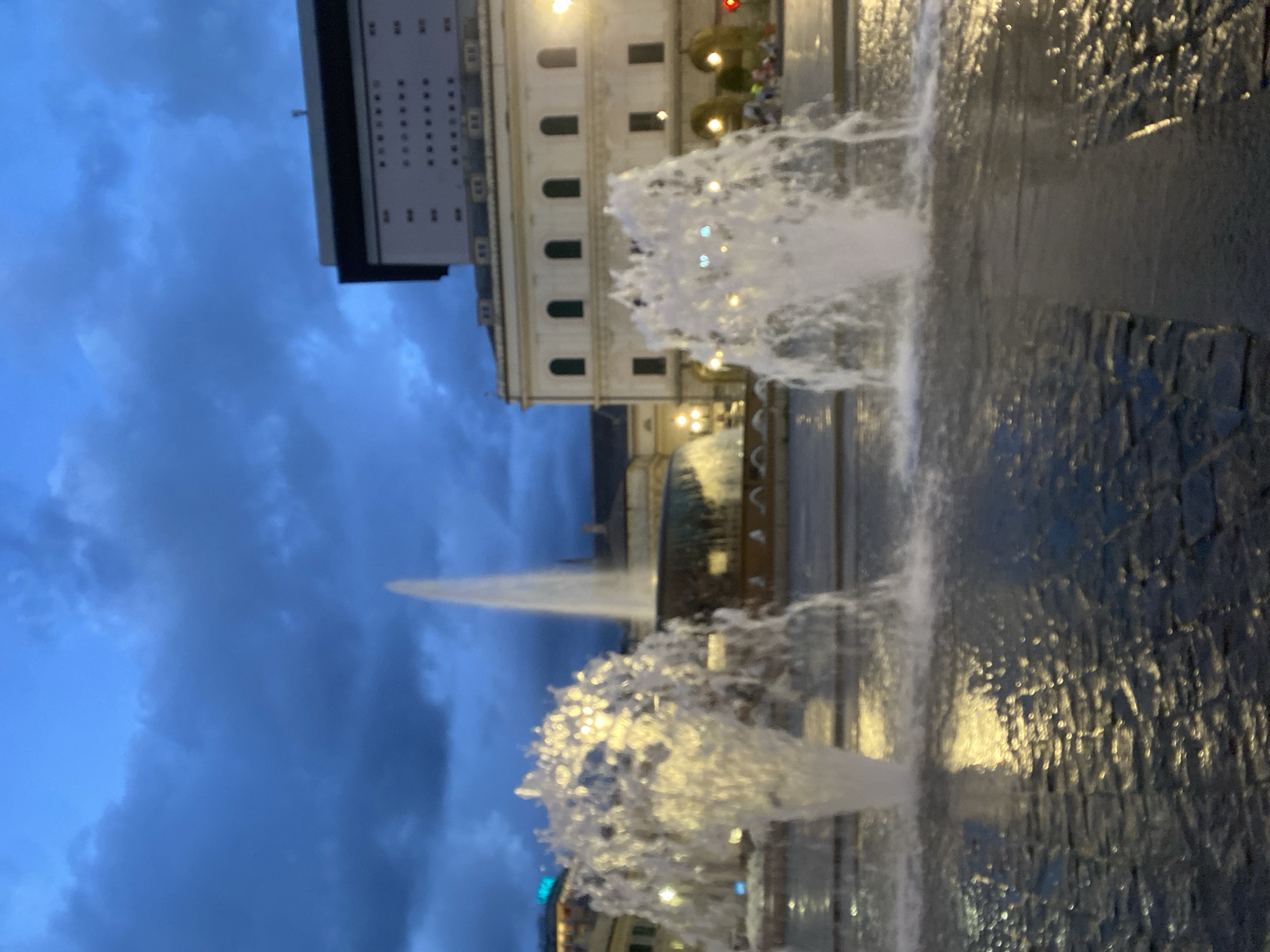 The image captures a public square in Genoa, Italy, at dusk or early evening. Two large fountains are prominently featured, with water jets spraying upwards and illuminated by artificial lights, casting reflections on the wet pavement. In the background, several buildings are visible, including a modern structure with many windows and an older, classical-style building. The sky is dark and cloudy, suggesting a potential for rain. There are a few indistinct figures of people in the background, moving about the square. The overall atmosphere is illuminated by artificial light sources.