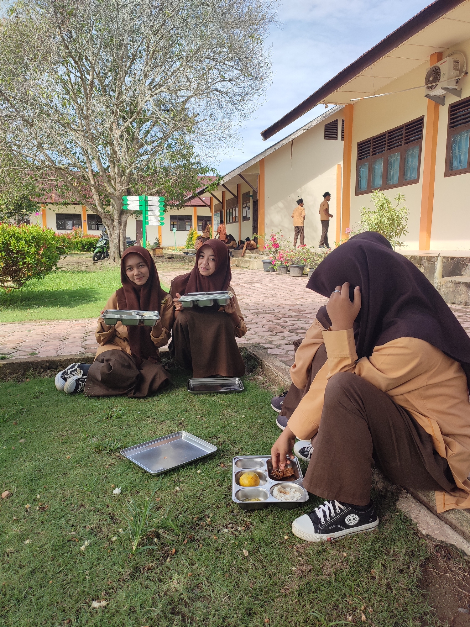 The image depicts an outdoor scene at a school campus in Blangriek, Indonesia, likely during a break or lunchtime. The bright daylight suggests it's daytime, under a clear or partly cloudy sky.
In the foreground, three female students, dressed in brown and light brown school uniforms with hijabs, are seated on a grassy area. The two students on the left and center are holding metal compartment trays and smiling at the camera. The third student, on the right, sits with her face covered by her hand, beside a metal tray containing a meal of rice, a whole mango, and other food items. An empty silver tray rests on the grass nearby. One pair of black sneakers with white laces, bearing the text "N.B." and "SPORT SHOE", is clearly visible.
The background features light-colored school buildings with brown roofs and window shutters. A large, deciduous tree with sparse foliage stands prominently to the left, and a green and white directional sign is visible beneath it. A paved area with bricks extends from the grass towards the buildings. Several male students, dressed in similar brown and light brown uniforms, are seen standing and sitting near the buildings in the mid-ground. Flowering plants in pots and a parked motorcycle are also visible in the distance. The overall scene suggests a casual moment of students enjoying their meals or resting outdoors.