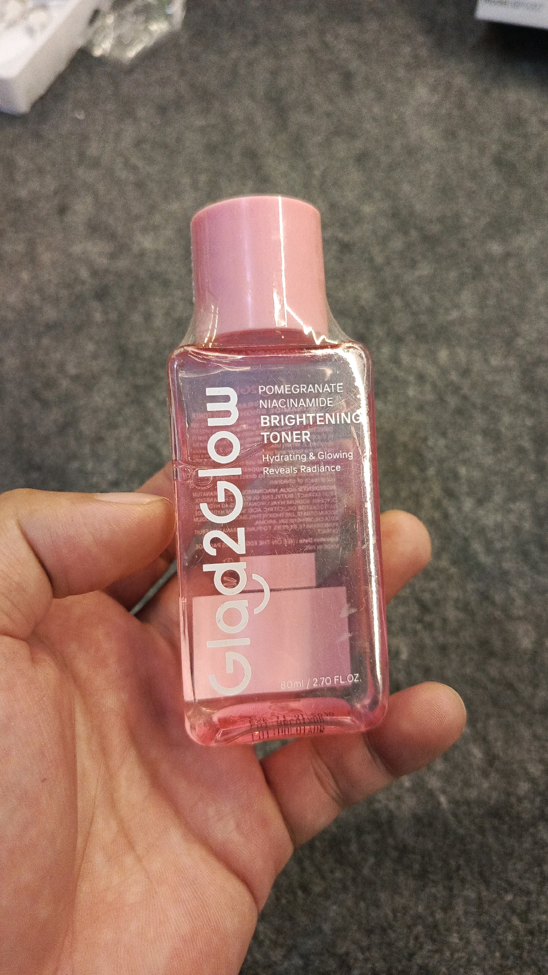A person's hand holds a small, clear plastic bottle with a pink cap, containing a pink liquid. The bottle is labeled with "Glow2Glow" in large white text and smaller text that reads "POMEGRANATE NIACINAMIDE BRIGHTENING TONER" and "Hydrating & Glowing Reveals Radiance." The bottle also indicates "80ml / 2.70 FL.OZ." The background is a textured, dark grey surface. The lighting suggests an indoor environment. No specific location in Babadan, Indonesia can be determined from the image.