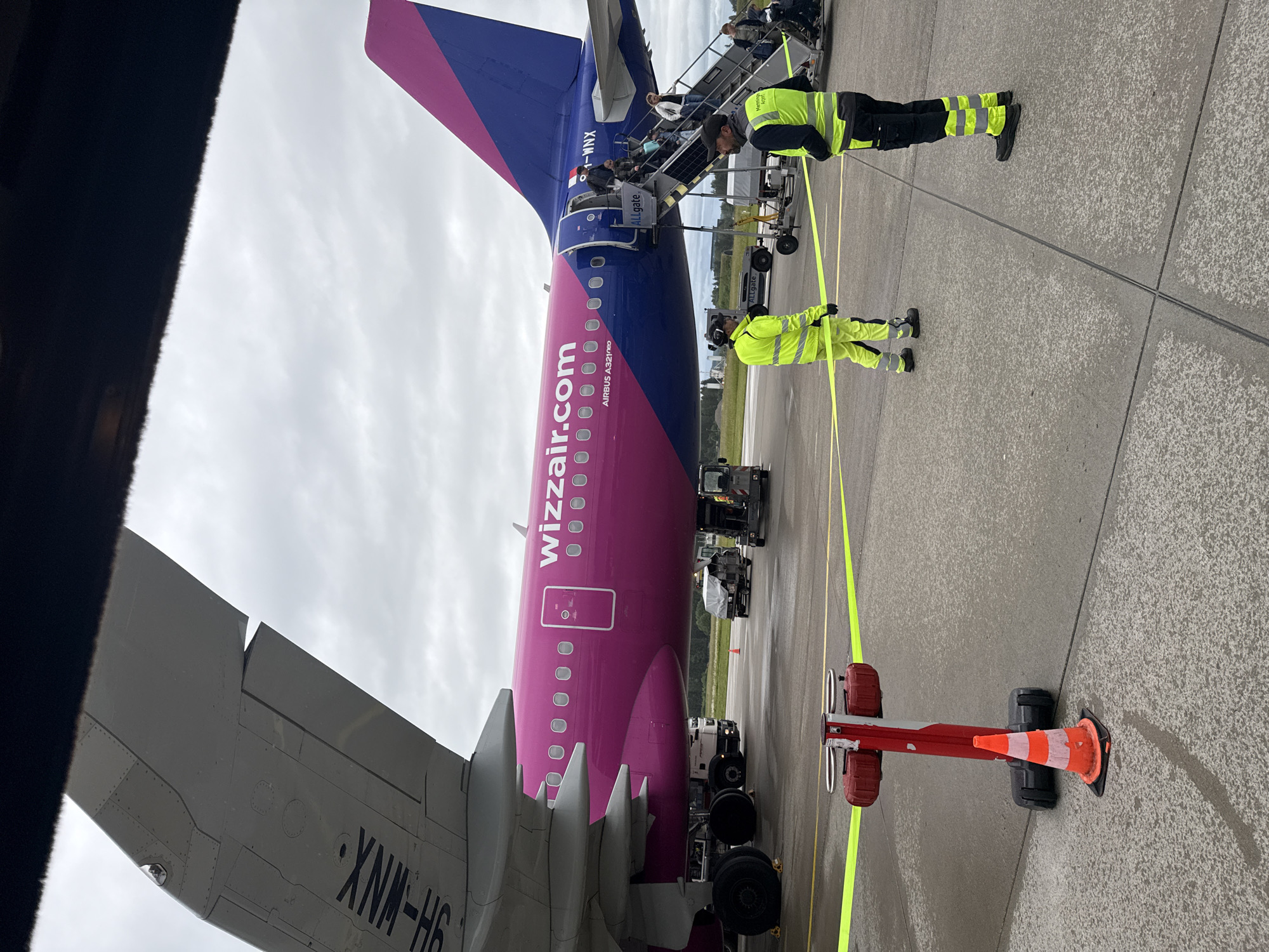 This image captures a Wizz Air Airbus A321neo aircraft on an airport tarmac, likely in Künersberg, Germany, under an overcast sky. The airplane, featuring a distinctive magenta and purple livery with "wizzair.com" emblazoned on its side, is in the process of boarding.
Passengers are seen ascending a mobile stairway positioned at the rear of the aircraft to enter the cabin. Two ground crew members, wearing high-visibility yellow-green uniforms, are present on the concrete tarmac. One crew member stands near the base of the stairs, while the other is further back, both positioned around a bright yellow safety tape that creates a perimeter. This tape extends from a red and white stanchion, next to which an orange traffic cone is placed.
Various pieces of ground support equipment are partially visible around and beneath the aircraft, including what appear to be baggage carts or tugs. The sky above is uniformly cloudy, suggesting an overcast day. In the background, beyond the tarmac, a line of green foliage is visible.
Visible text on the aircraft includes "wizzair.com" and "AIRBUS A321neo" on the fuselage, and the aircraft registration "HA-MNX" on the tail fin. The mobile stairs are marked "ALLgate," and "XNM-H6" is visible on the underside of a wing. Part of the text "Allgate Airport" is also discernible on the vest of one of the ground crew members. A dark, out-of-focus element in the foreground on the left suggests a window frame or part of a building.