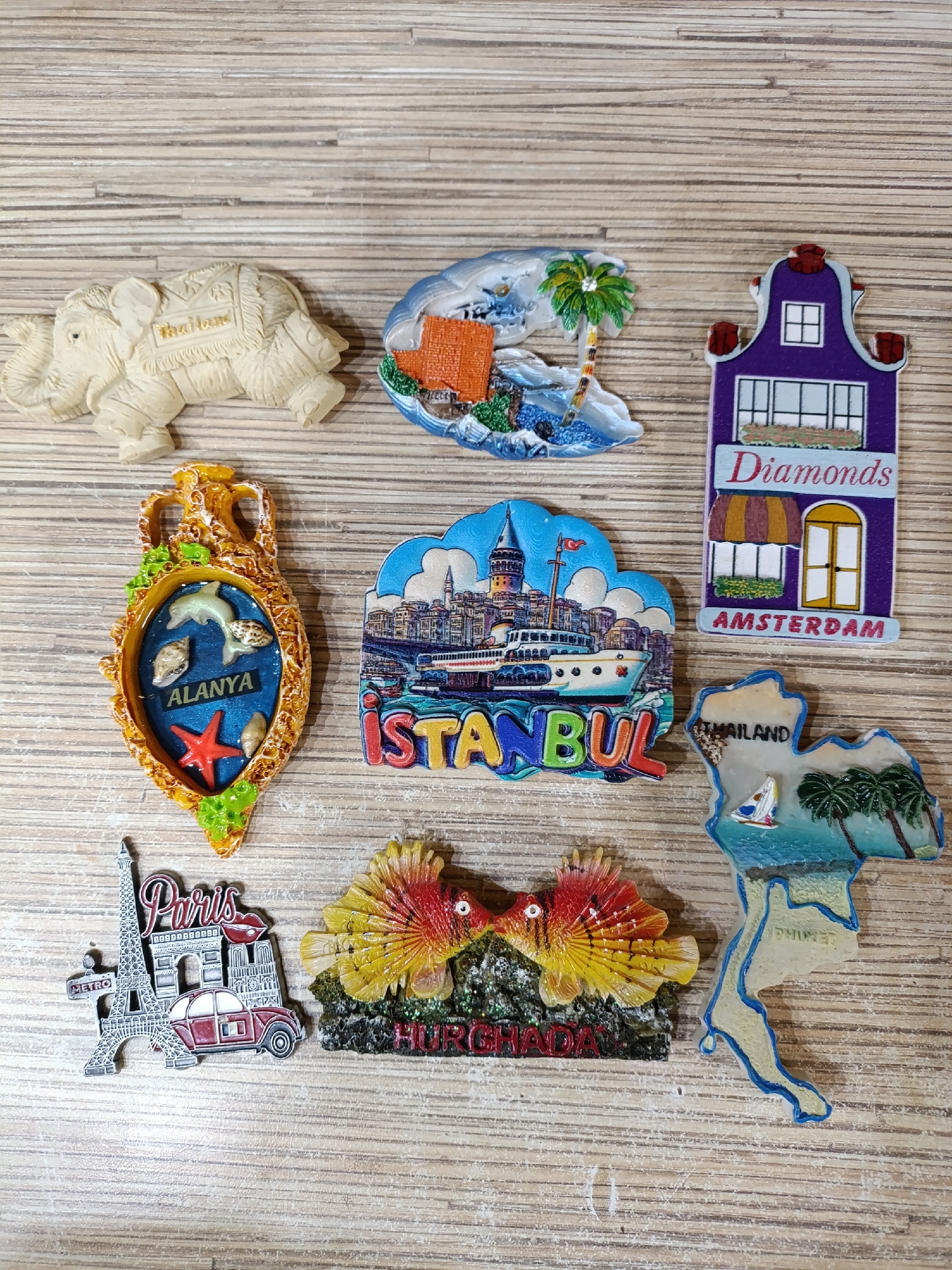 The image shows a collection of souvenir magnets arranged on a wooden surface. The magnets are from various international locations including Thailand, Amsterdam, Alanya, Istanbul, Paris, Hurghada, and Phuket. The magnets feature landmarks, cityscapes, and cultural symbols associated with each location. There are no people present. The wooden surface appears to be a tabletop or other flat surface. The lighting is bright and even, allowing for clear visibility of the magnets. The location is unknown, and there is no indication of the city of Chaikovsky, Russia.